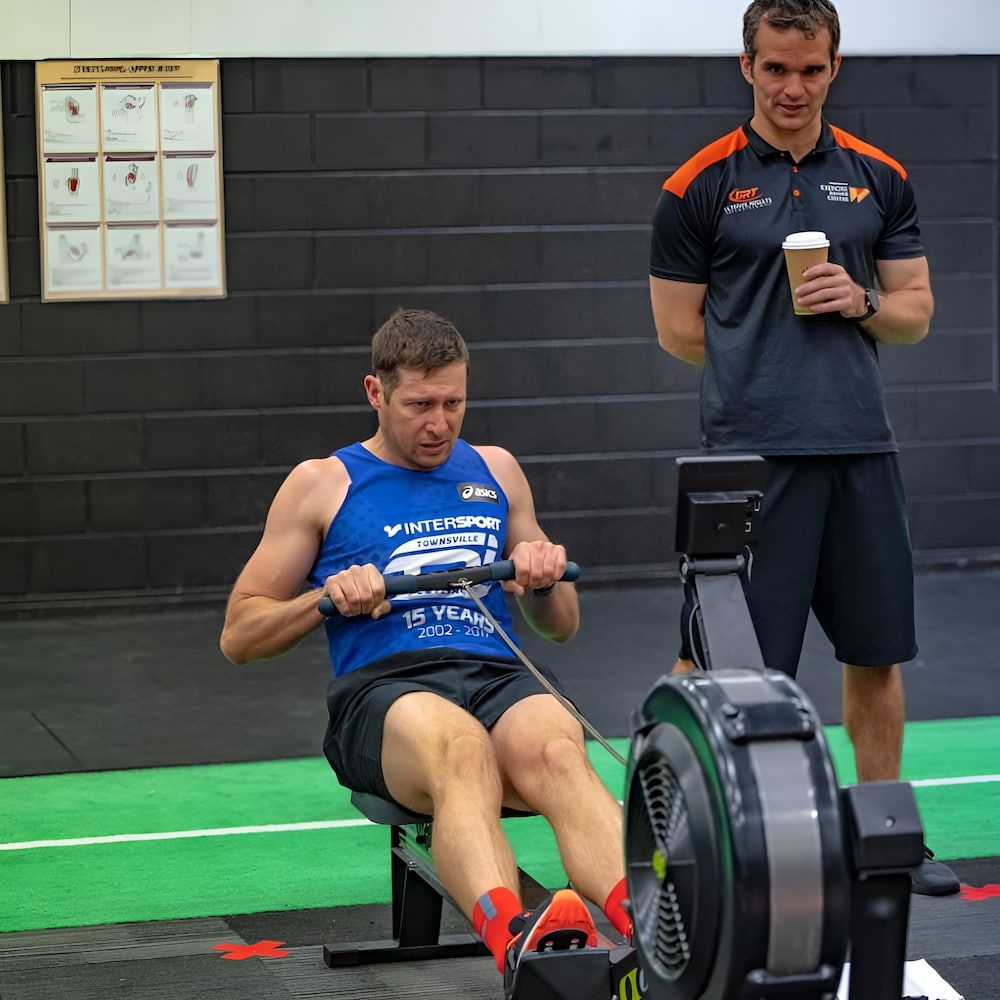 A Man Is Using A Rowing Machine While Another Man Watches — Ultimate Results Training in Aitkenvale, QLD