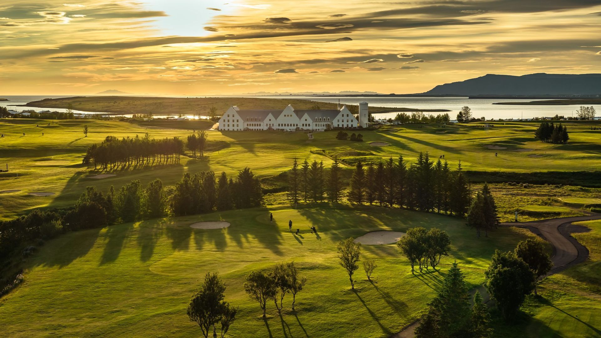 Sólbjartur golfvöllur með löngum skuggum, trjám, krókóttum stíg og hvítum byggingum nálægt vatni við sólsetur