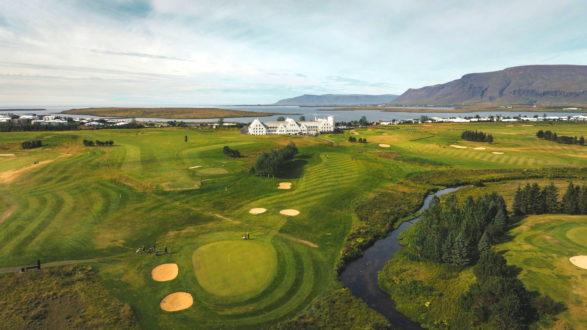 Golfvöllur með sandbyrgjum við á, hvítum byggingum og fjöllum undir skýjuðum himni