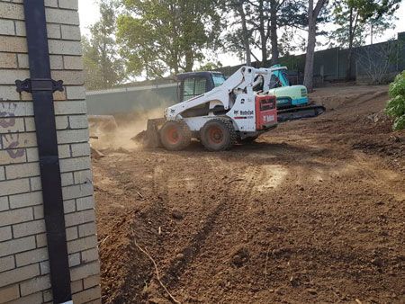 A bobcat is driving through a dirt field next to a brick building.