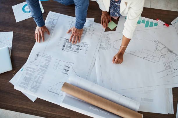 Two people reviewing architectural blueprints spread on a table. Beige, white, and blue plans with graphs and notes, viewed from above.