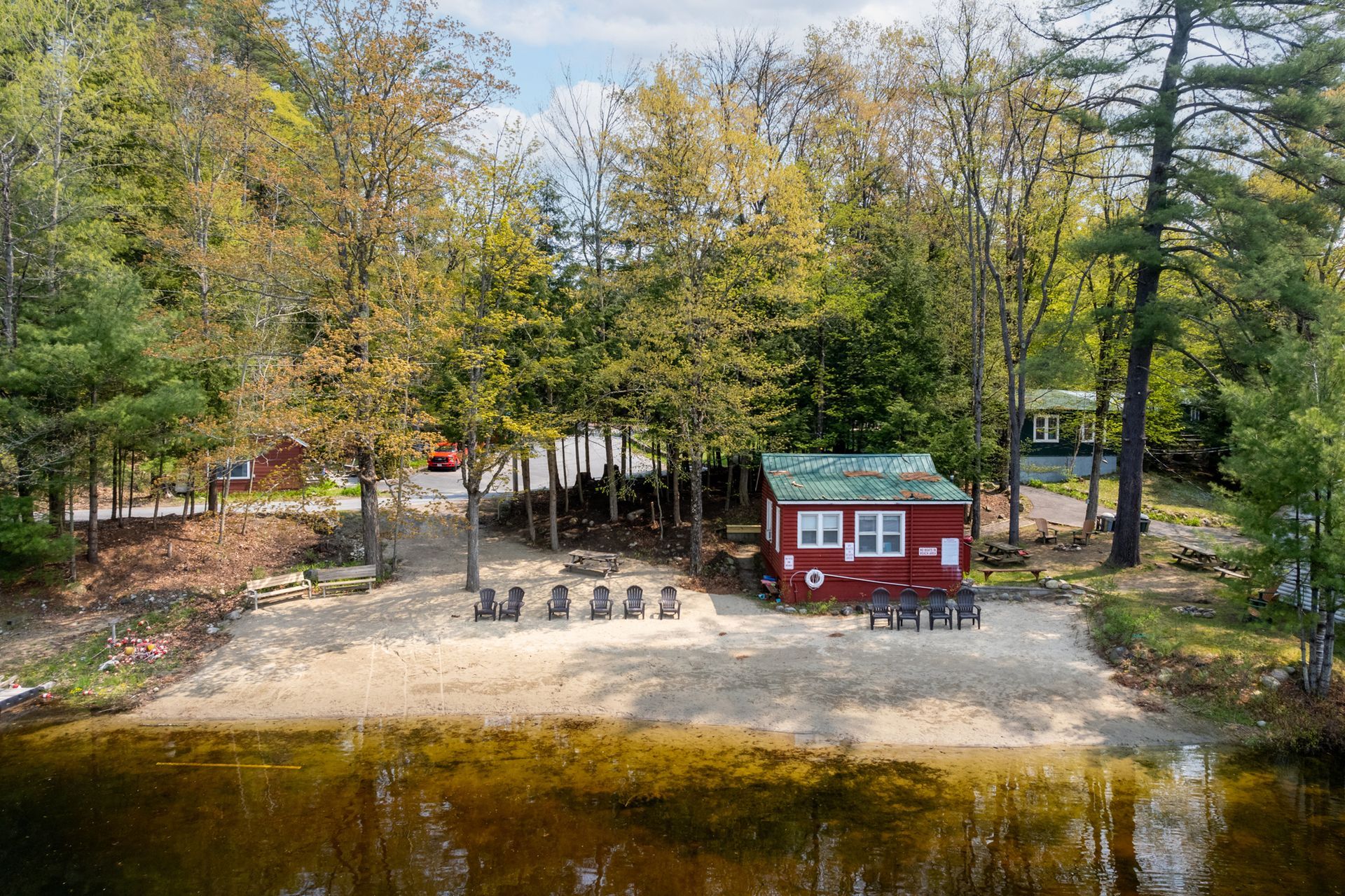 Red cabin on sandy shore of a lake, with trees in the background and chairs on the beach.