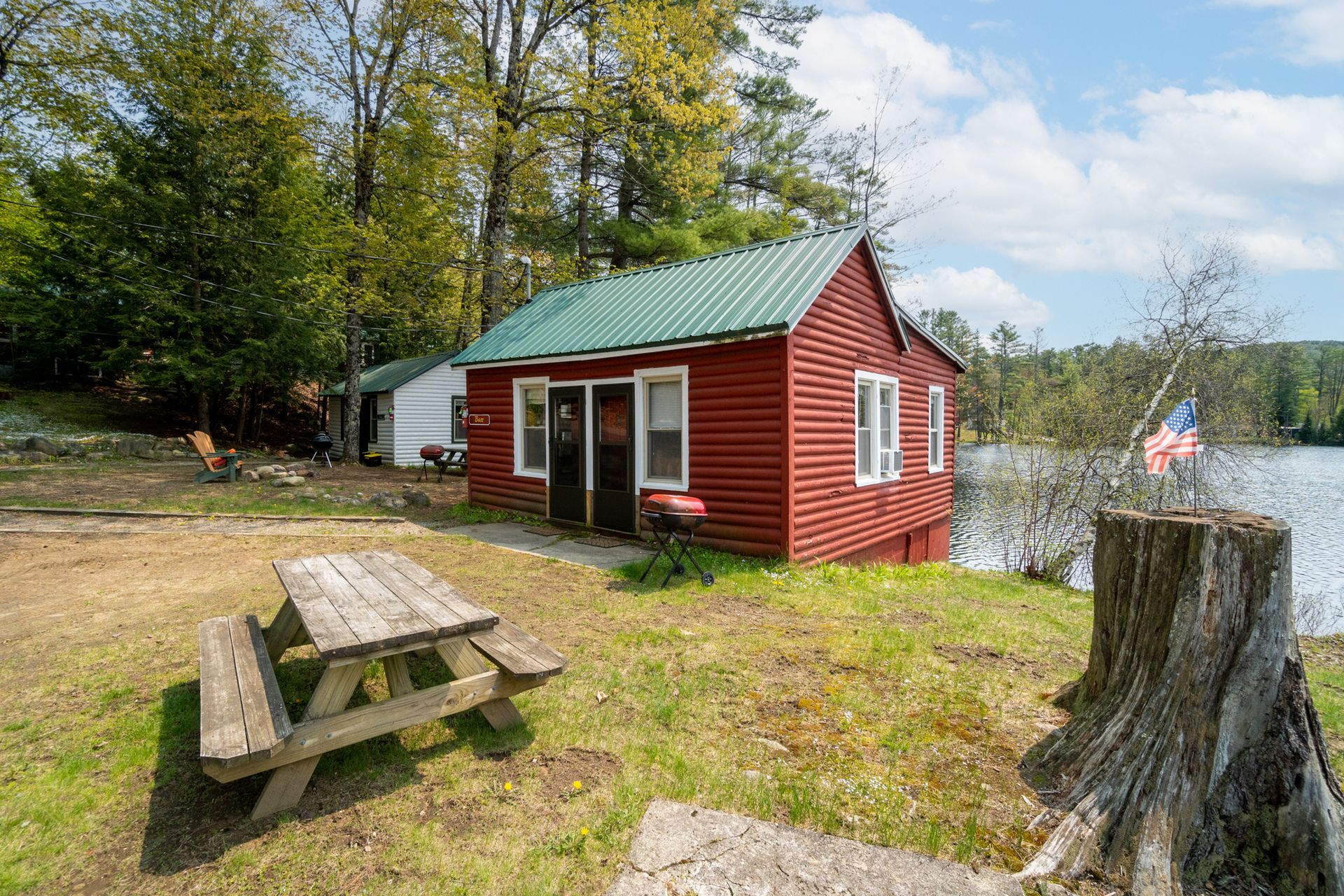 Red cabin with green roof by a lake, picnic table in front.