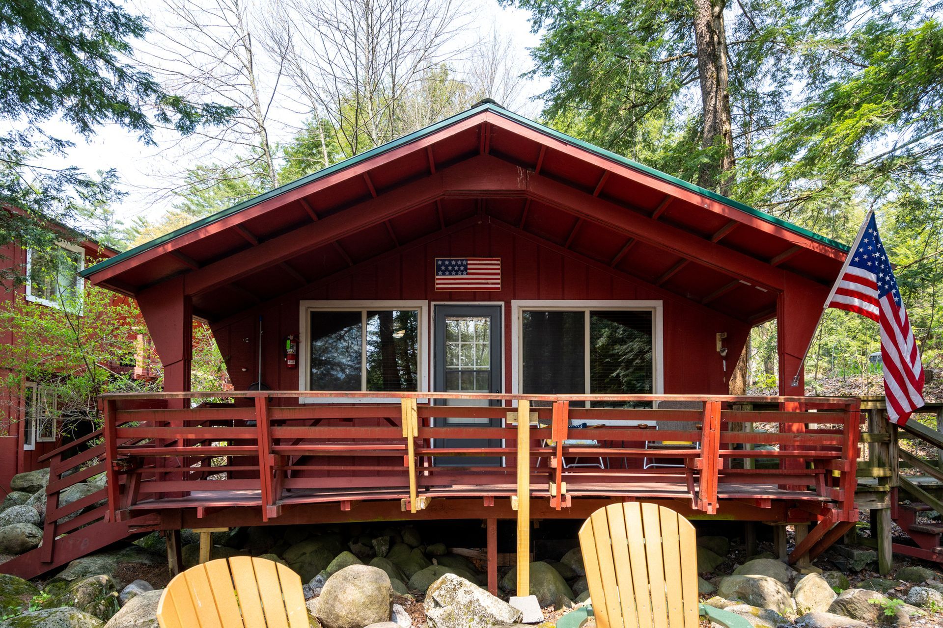 Red cabin with wooden porch and American flag; surrounded by trees and rocks.