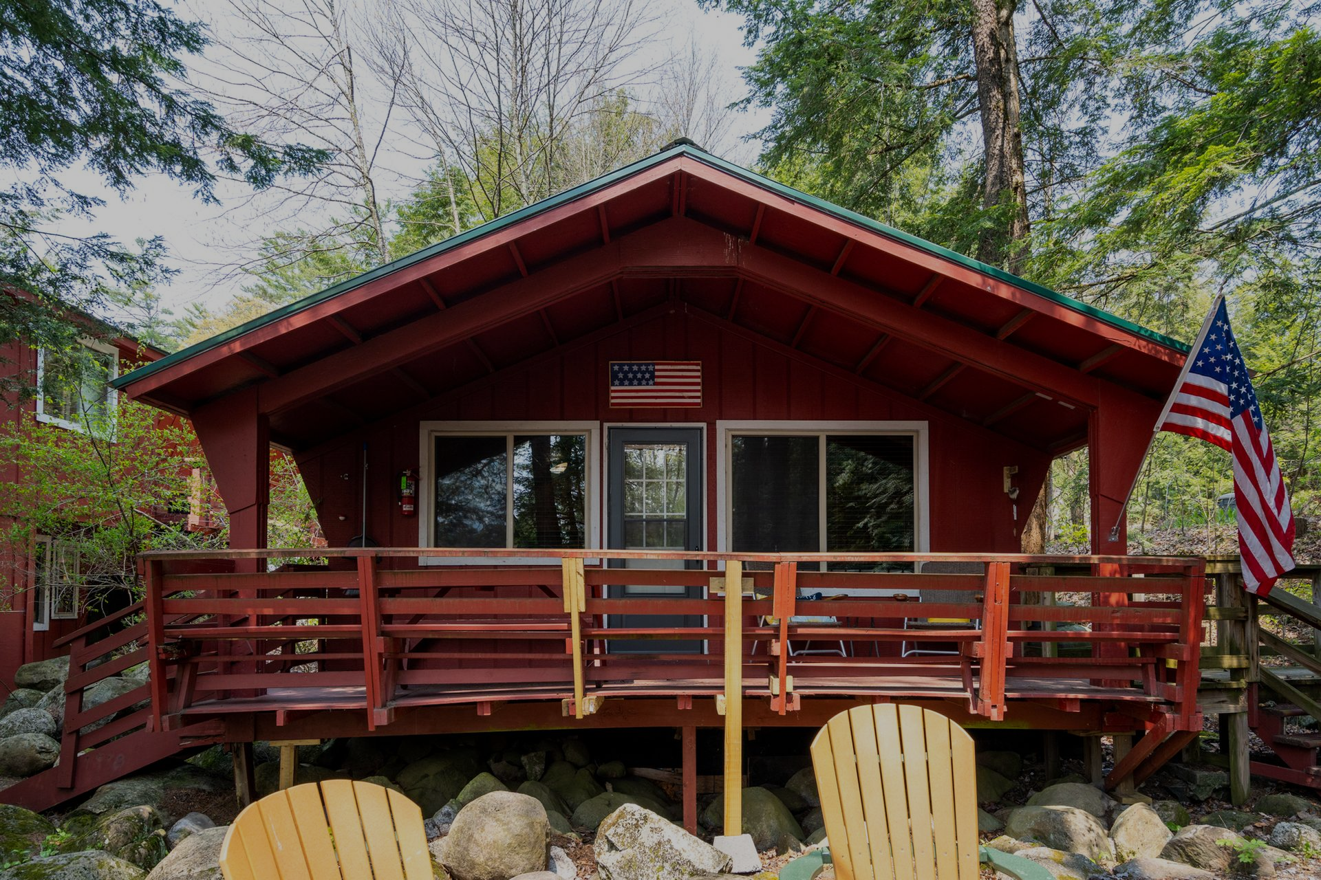 Red cabin with porch and American flag, nestled among trees.