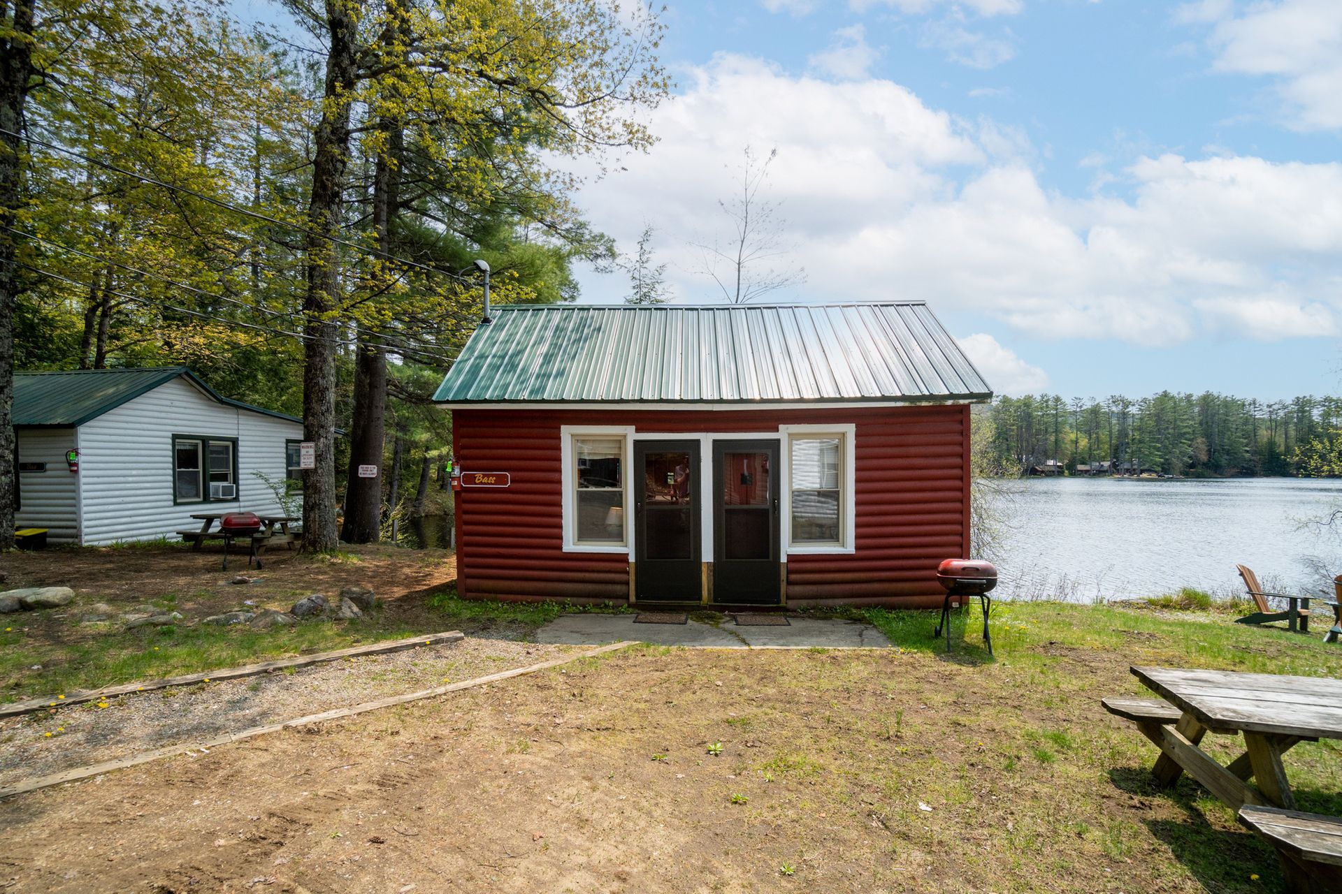 Red and white cabins by a lake, trees in background, sunny day. Picnic table and grill in the foreground.