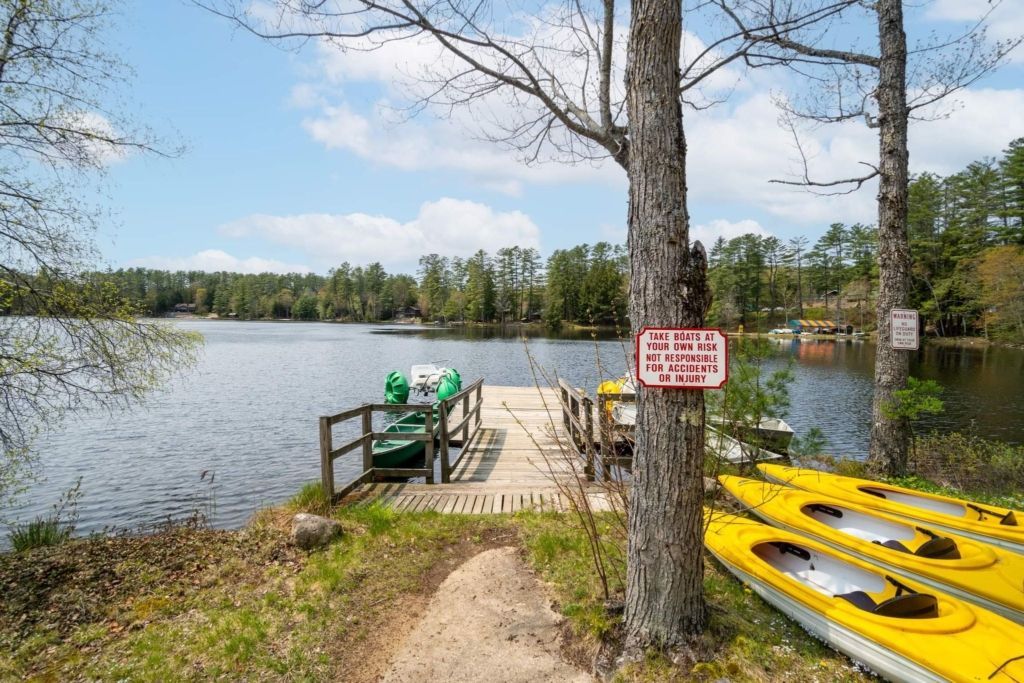 Dock on a lake with kayaks, boats, and a sign. Trees surround the water under a blue sky.