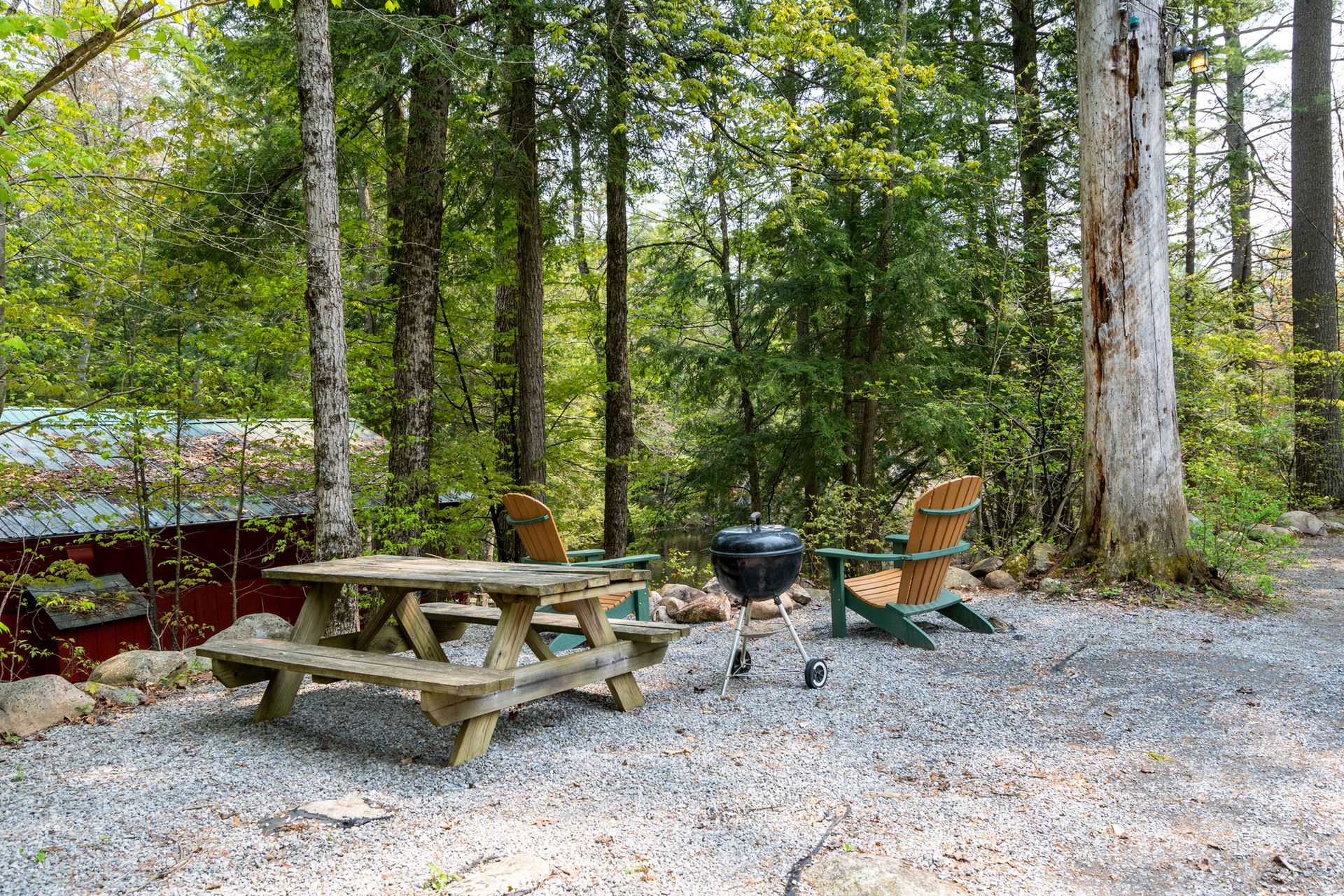 Picnic table, chairs, and grill in a gravel clearing surrounded by trees, possibly a campsite.