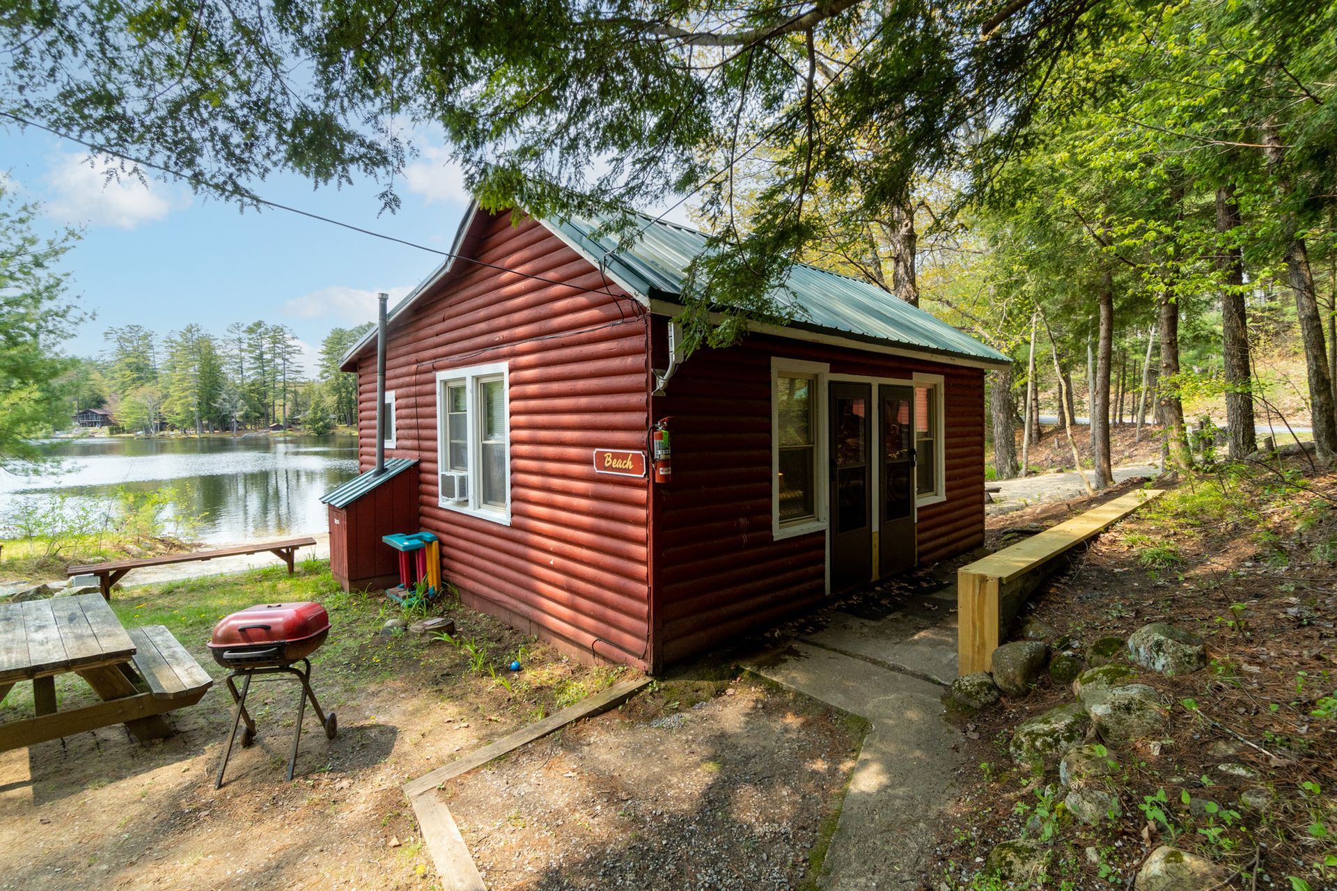 Red cabin by a lake with a picnic table and grill.
