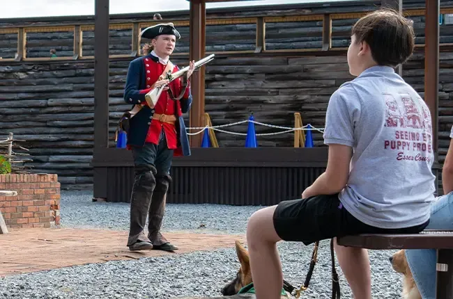 Person in colonial attire demonstrates a musket to a seated person outdoors, with wood and brick backdrop.