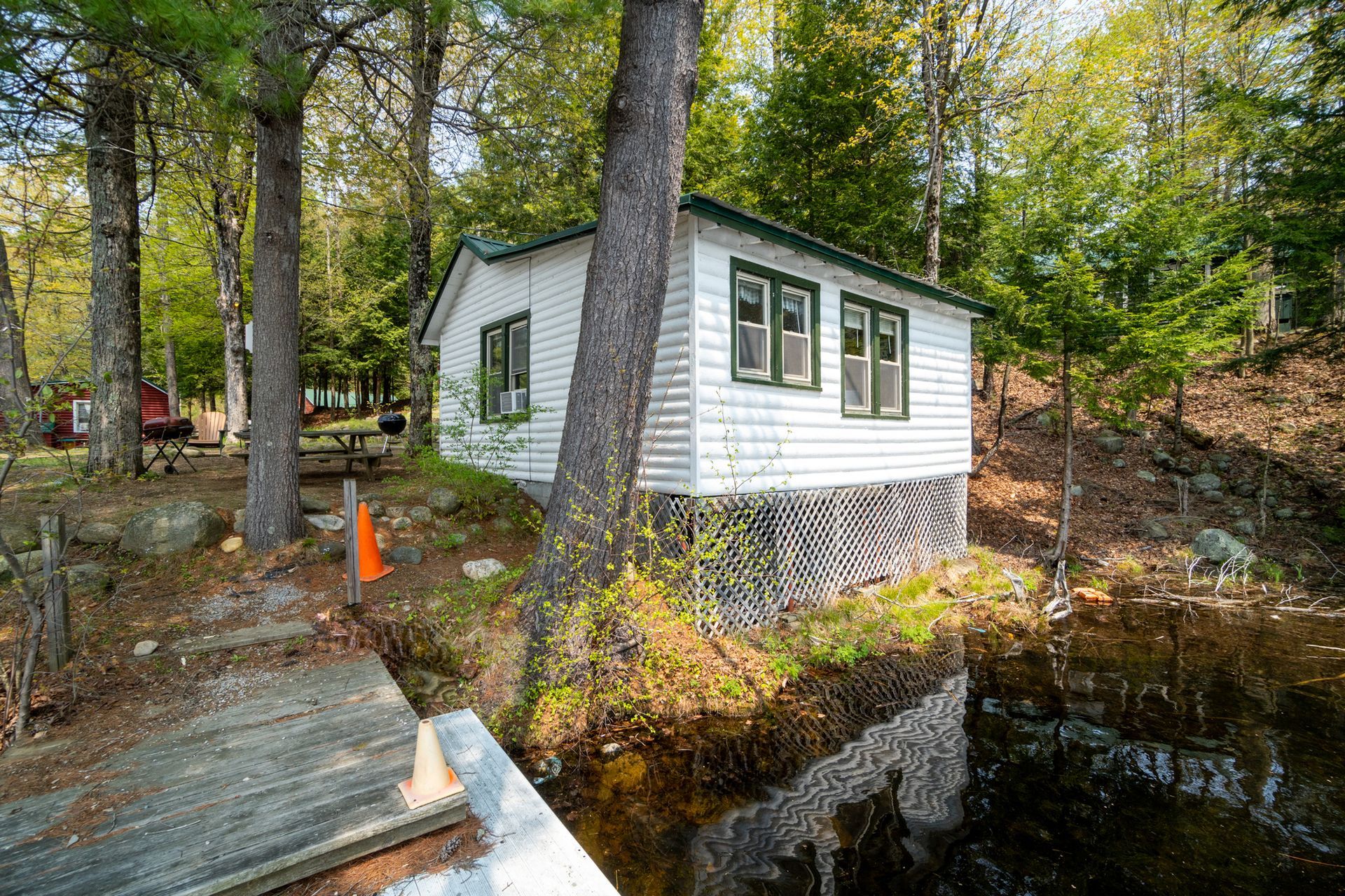 Small white cabin on the edge of water, with green trim, set amongst trees and a weathered dock.