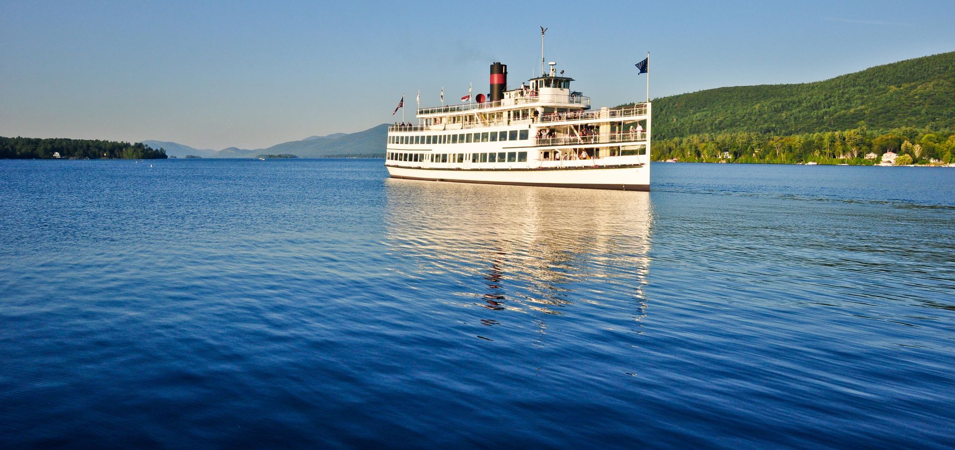 A large white steamboat sails on a blue lake; green mountains and clear sky in the background.