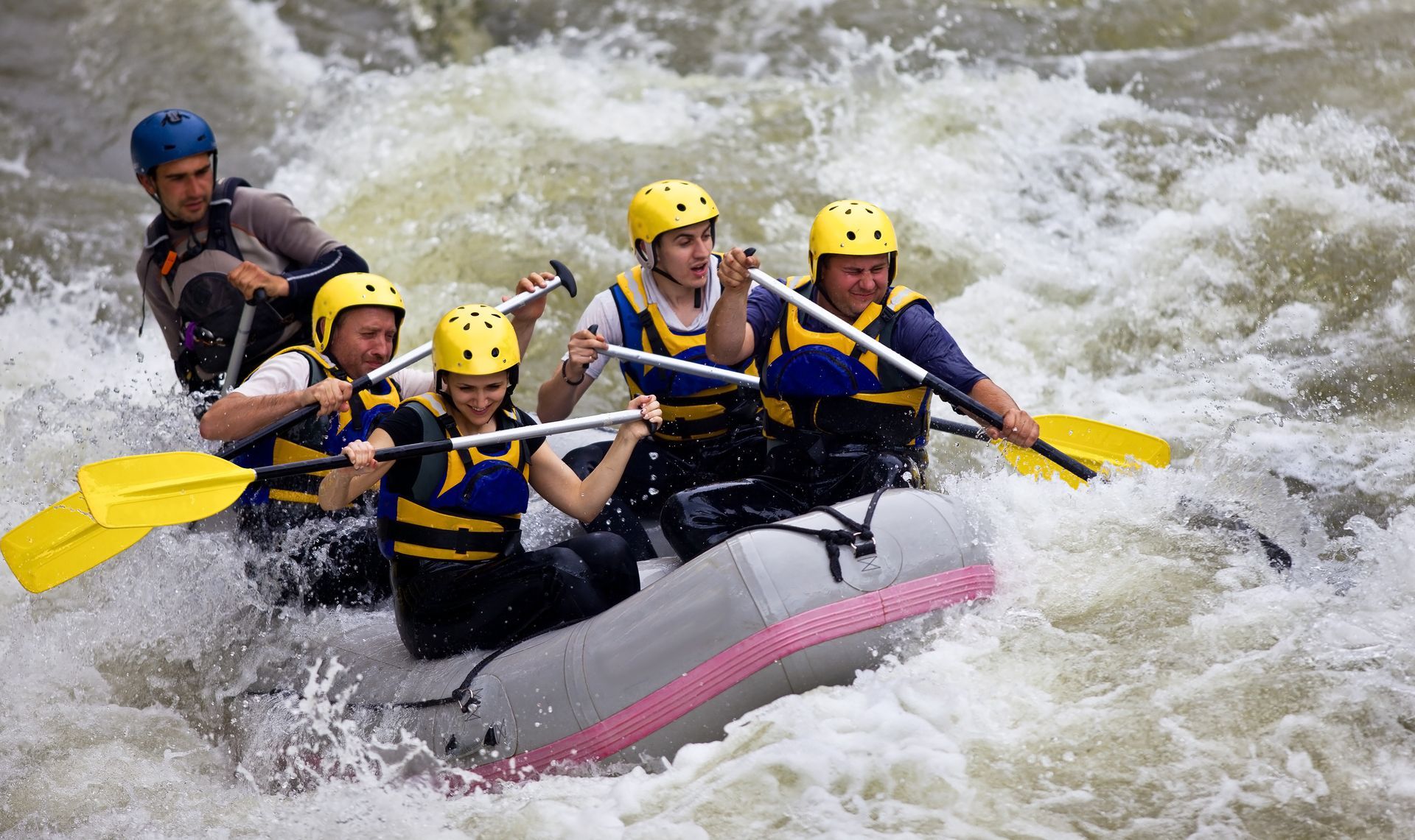 People white water rafting through churning, foamy water. They wear helmets and life vests, paddling.