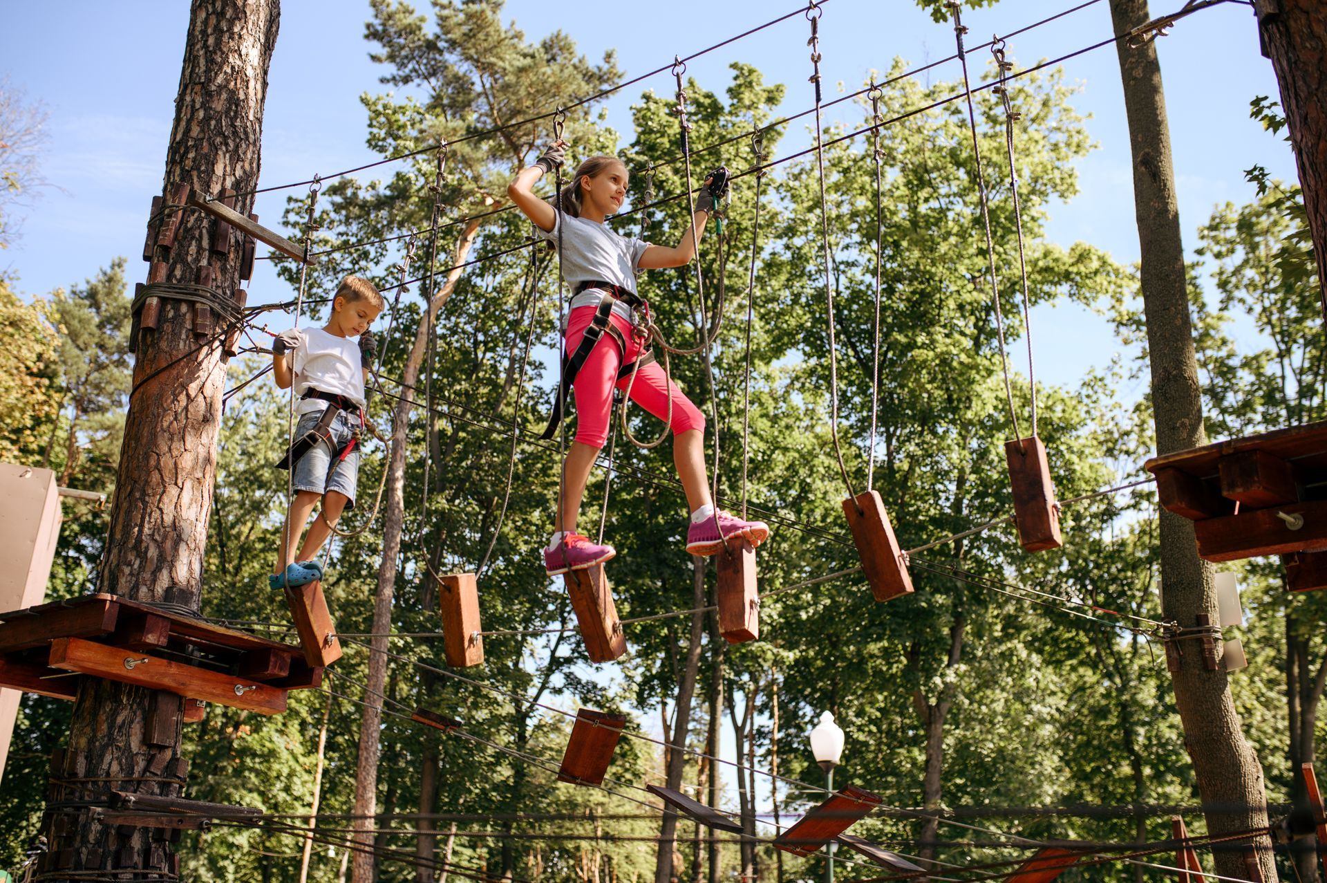 Two children on an elevated ropes course, balancing on wooden planks. Trees and blue sky in background.