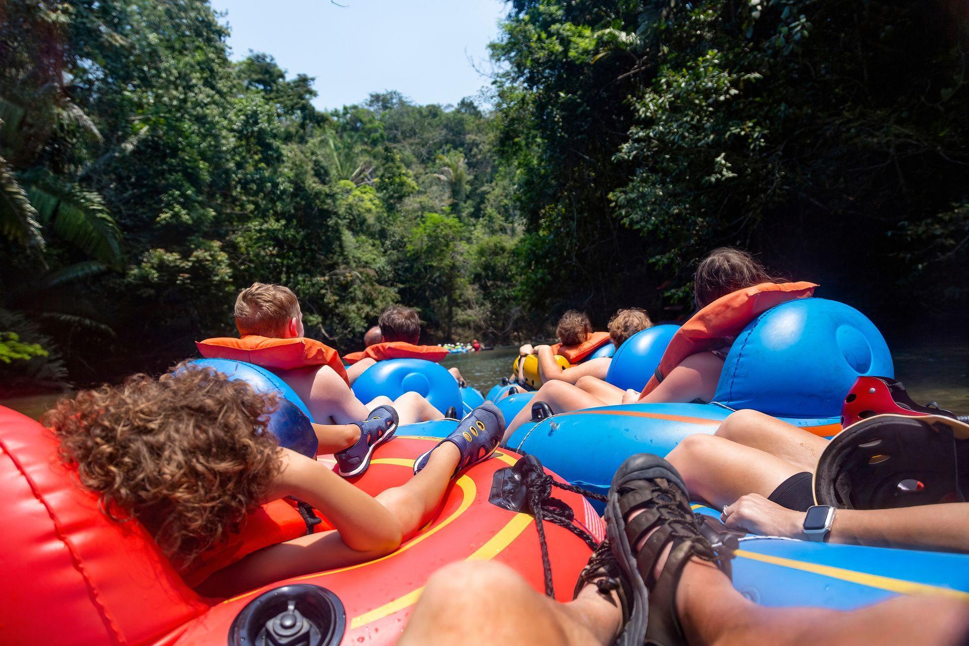 People tubing down a river, wearing life vests, surrounded by green trees.