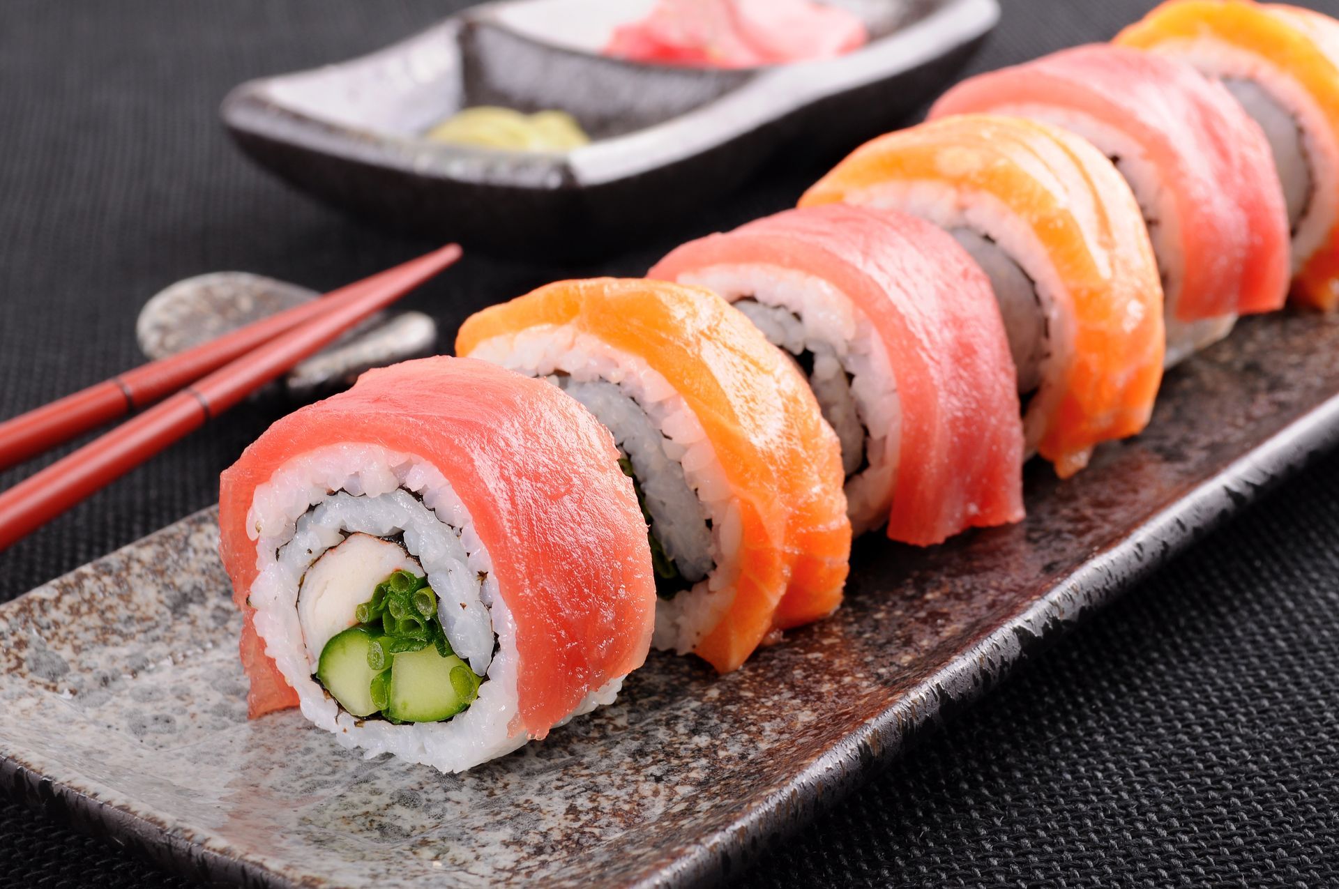 Sushi roll pieces with colorful fish, on a rectangular plate, with chopsticks and dipping sauces.