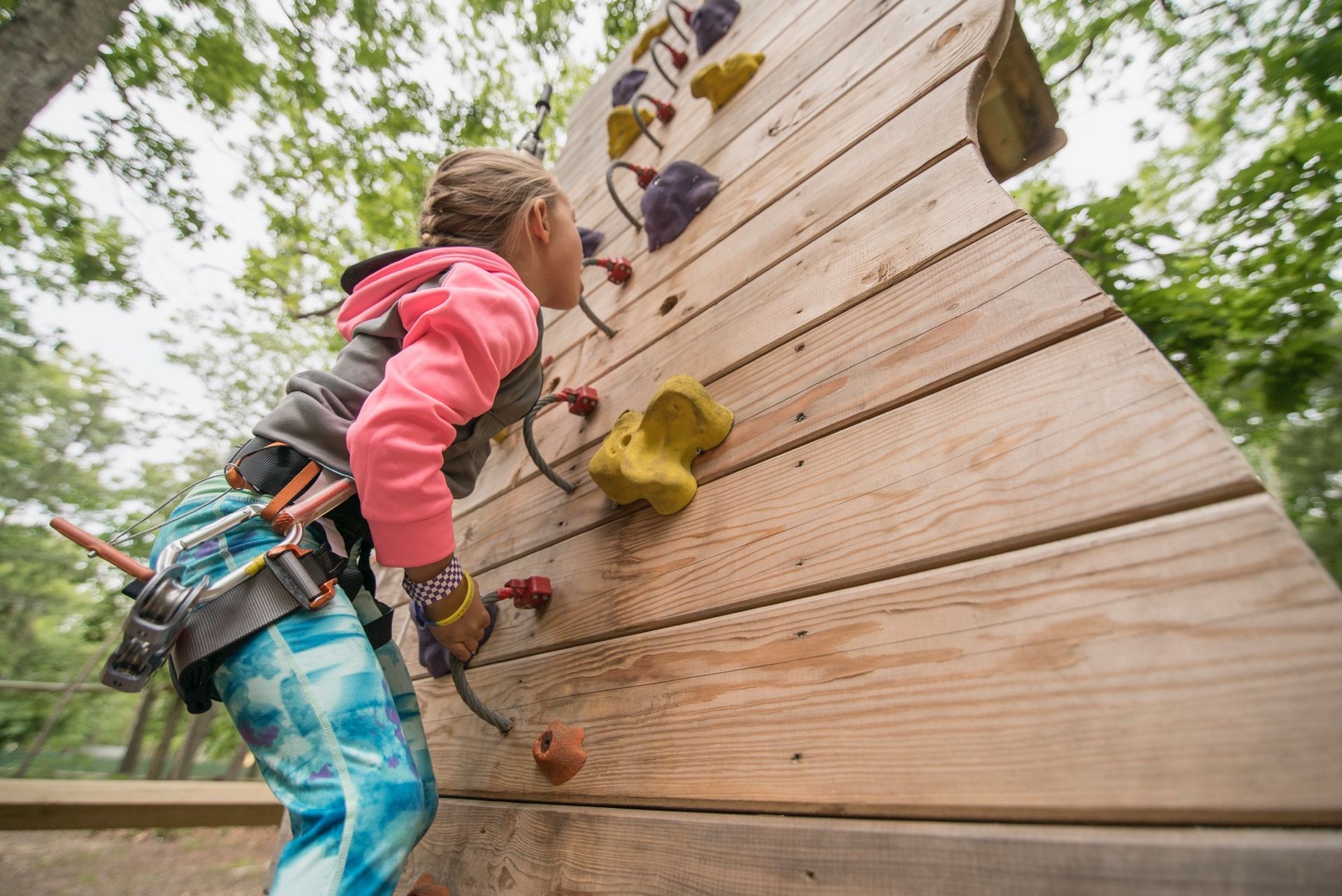 Girl climbing a wooden wall with colored handholds, wearing safety harness in an outdoor setting.