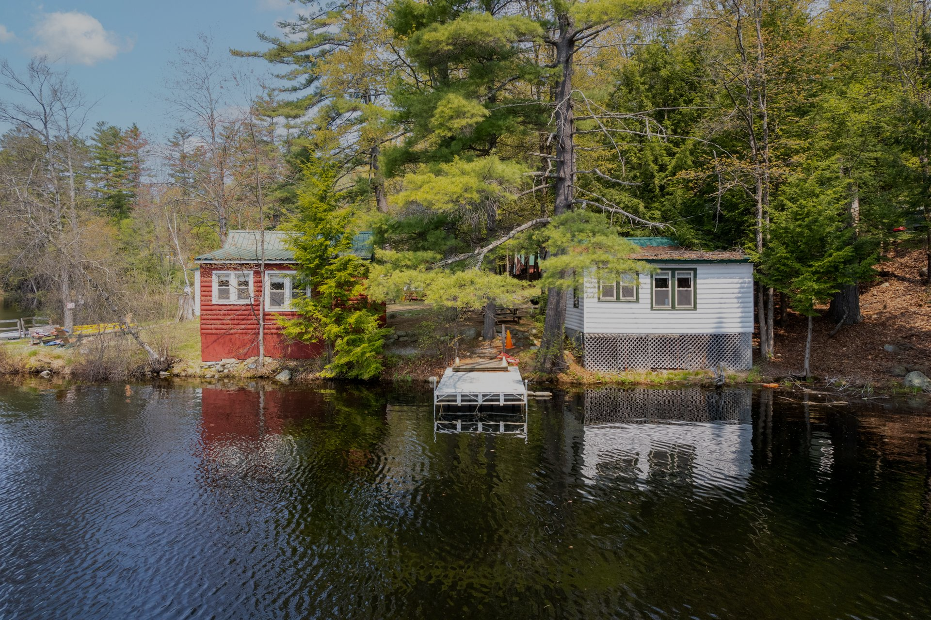 Two small, weathered buildings on a lake's edge. One red, one white, with reflections in dark water.