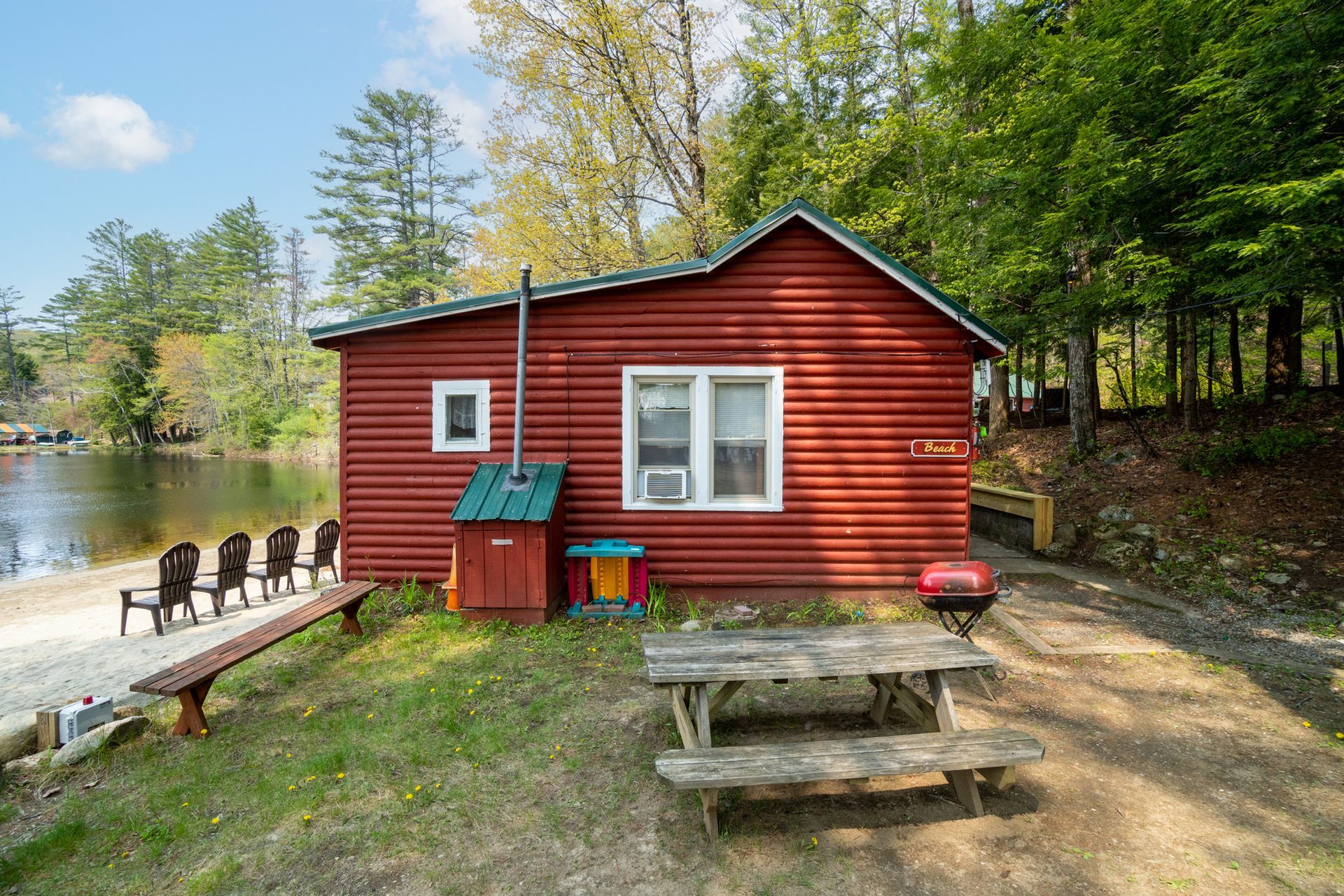 Red cabin by a lake with picnic table and chairs on a dock.
