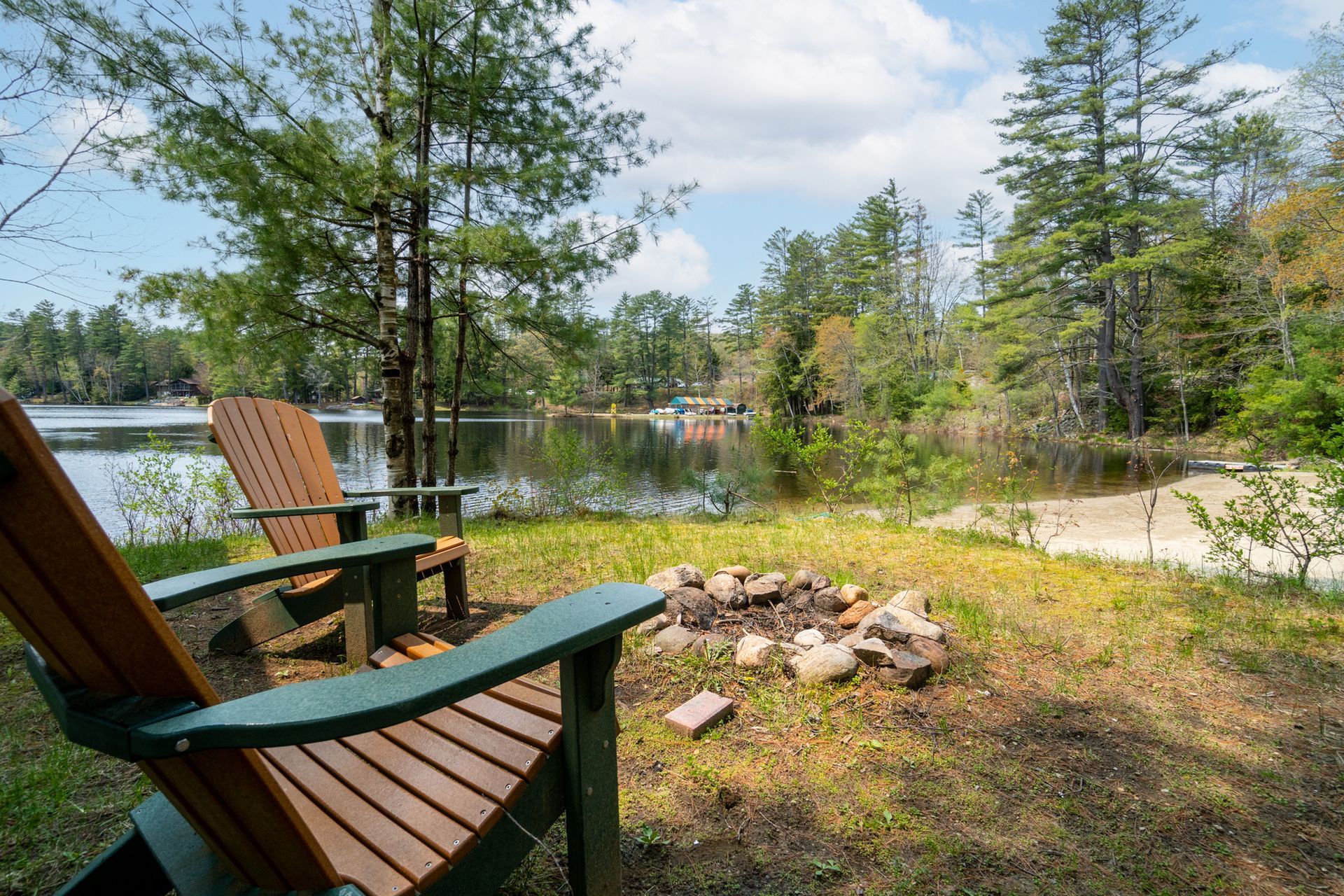 Two wooden chairs face a lake next to a small fire pit, with trees in the background.