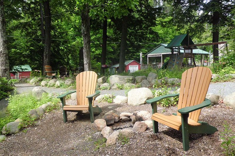 Two wooden Adirondack chairs face a stone fire pit in a wooded area. Playground visible behind.