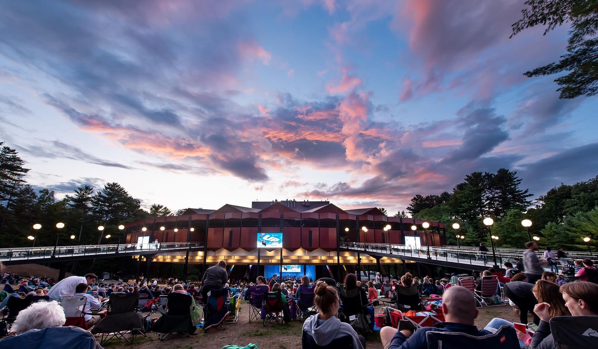 People seated outdoors watching a performance on a stage at dusk; colorful sky overhead.