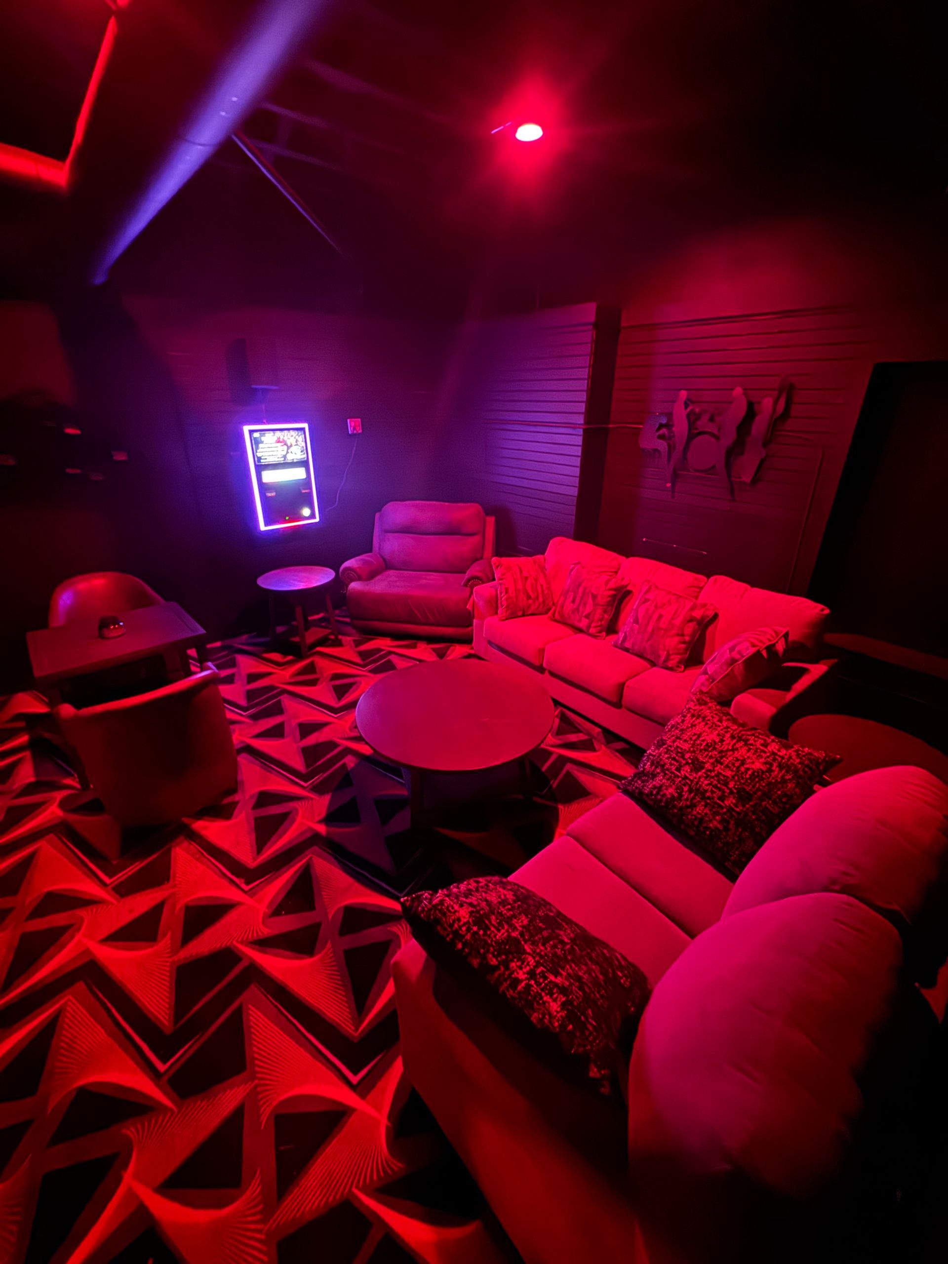 A dimly lit room with red lights, featuring a seating area with a couch, chairs, and a slot machine.