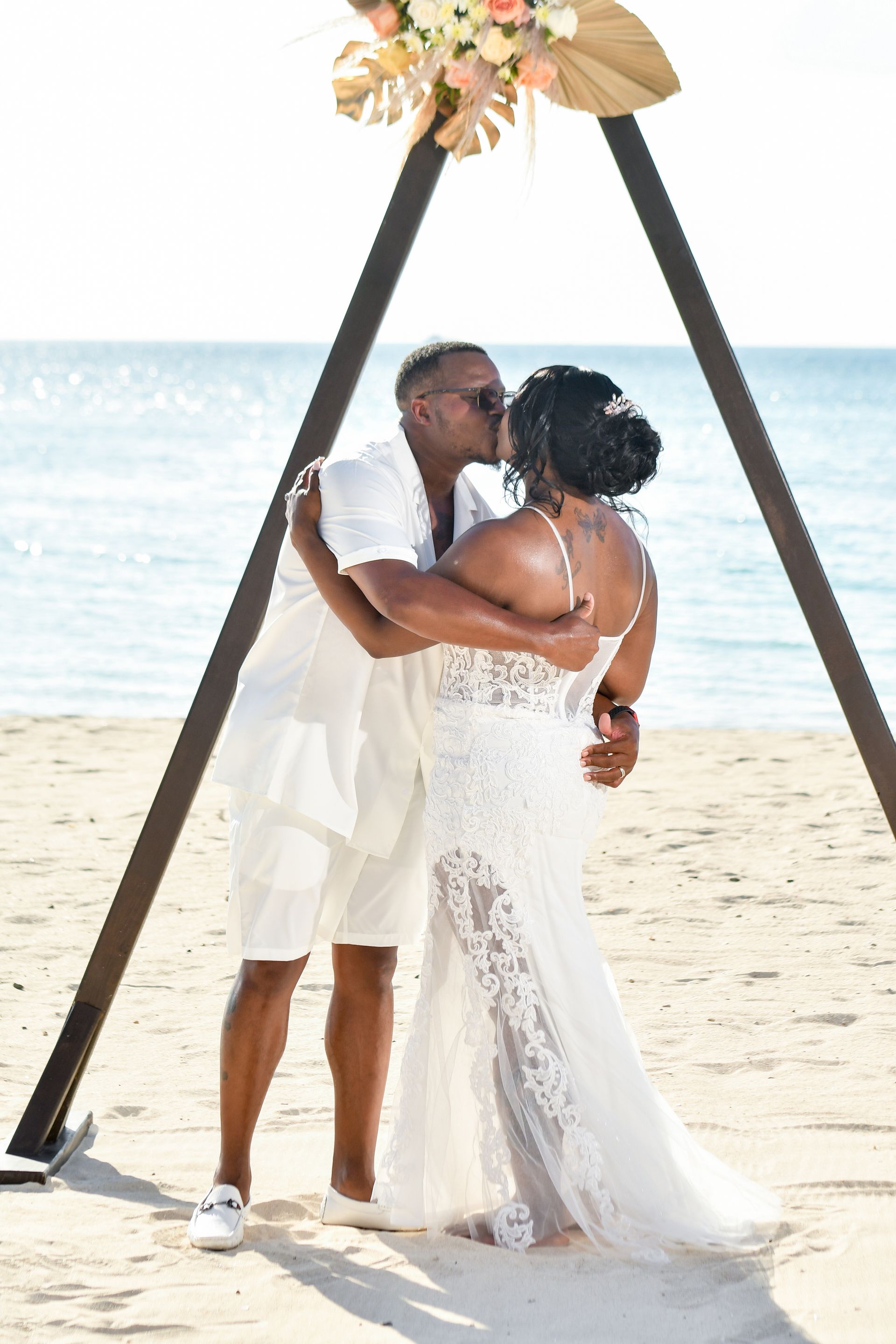 A bride and groom are kissing under a wooden arch on the beach.