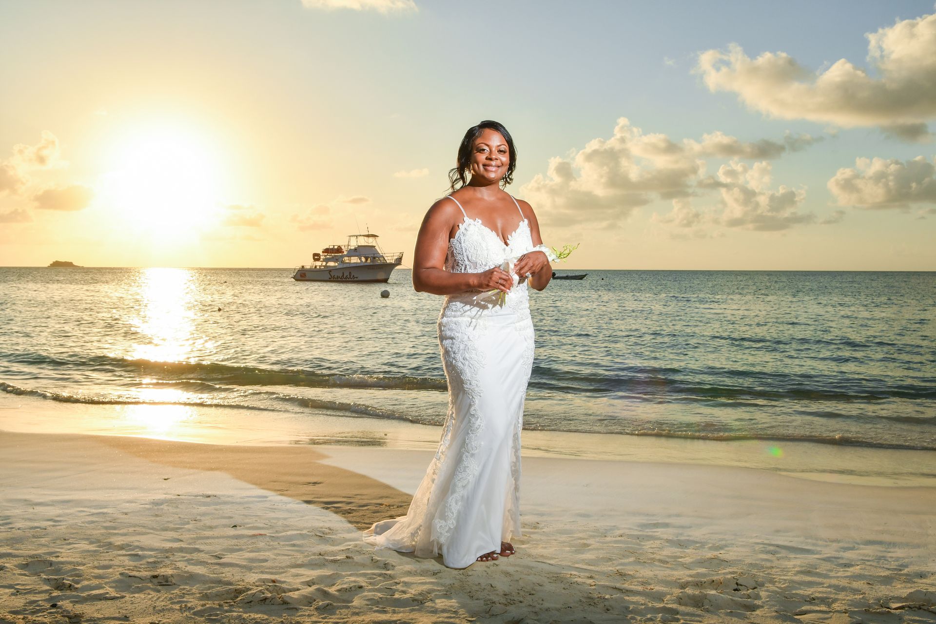 A woman in a wedding dress is standing on a beach with a boat in the background.
