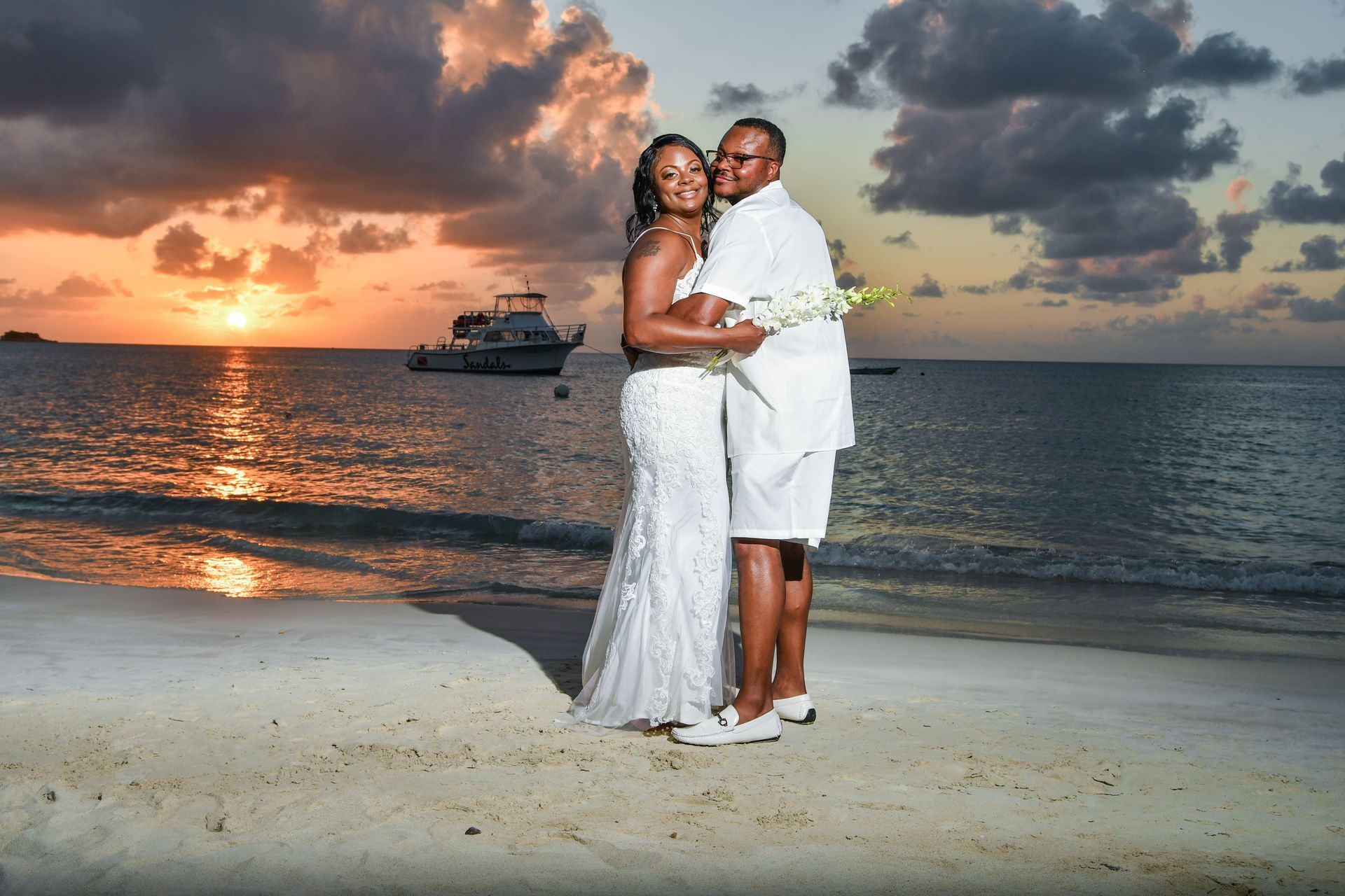 A bride and groom are posing for a picture on the beach at sunset.