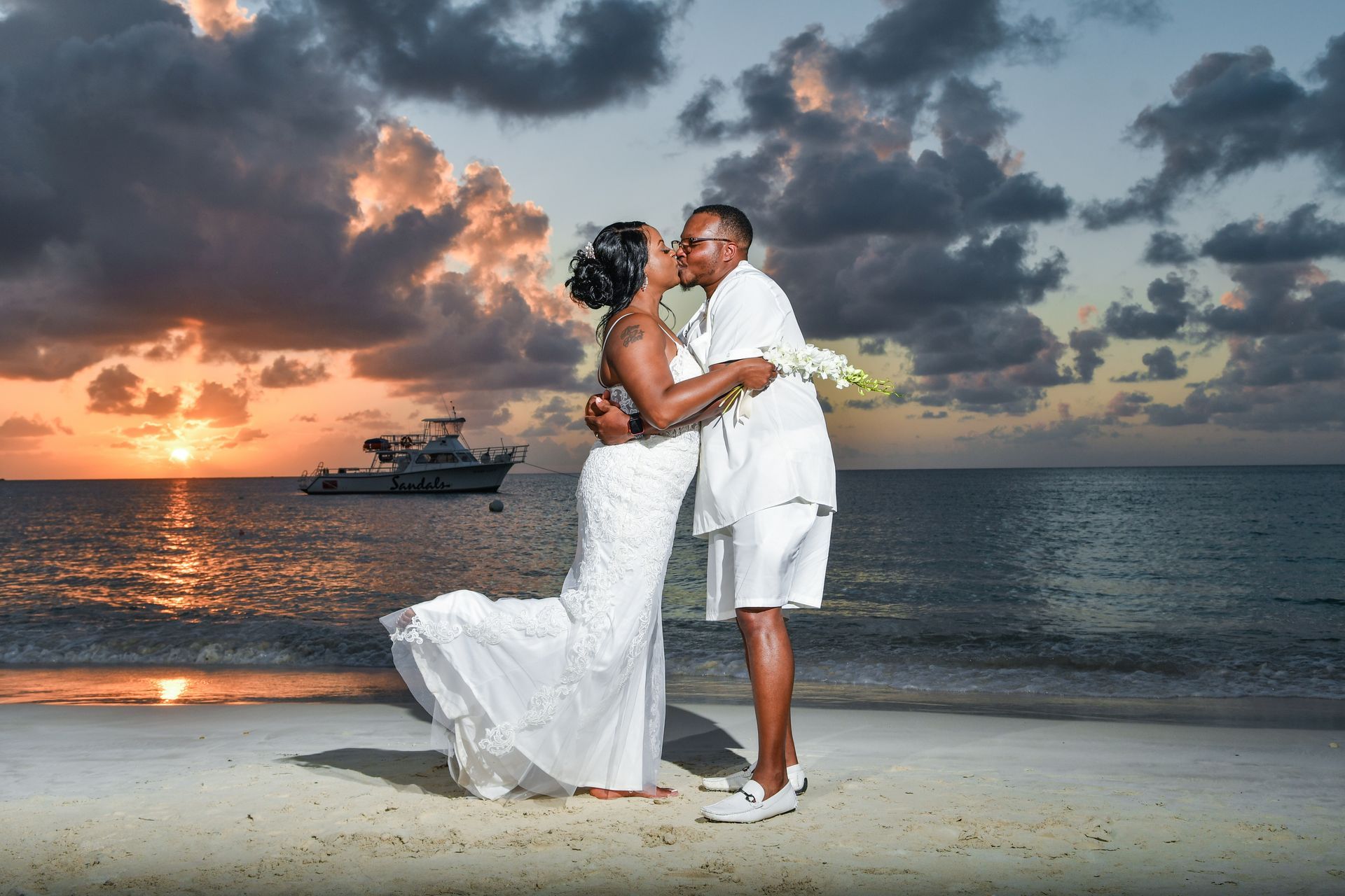 A bride and groom are kissing on the beach at sunset.