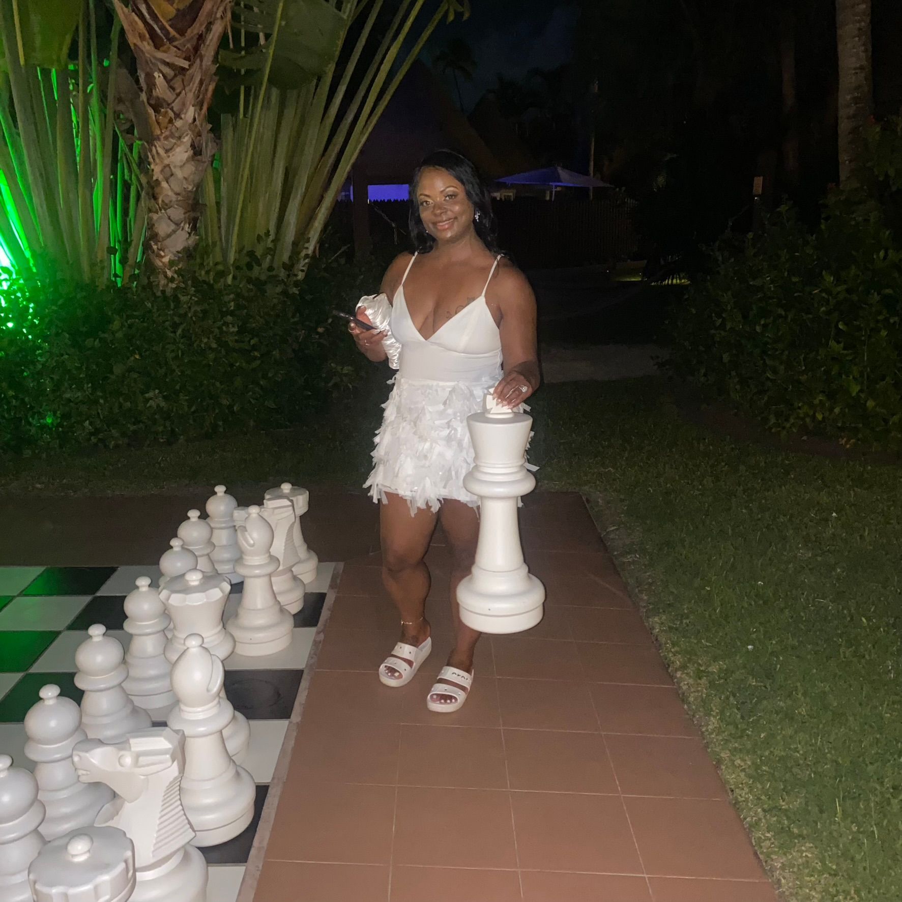 A woman in a white dress is standing next to a giant chess board.