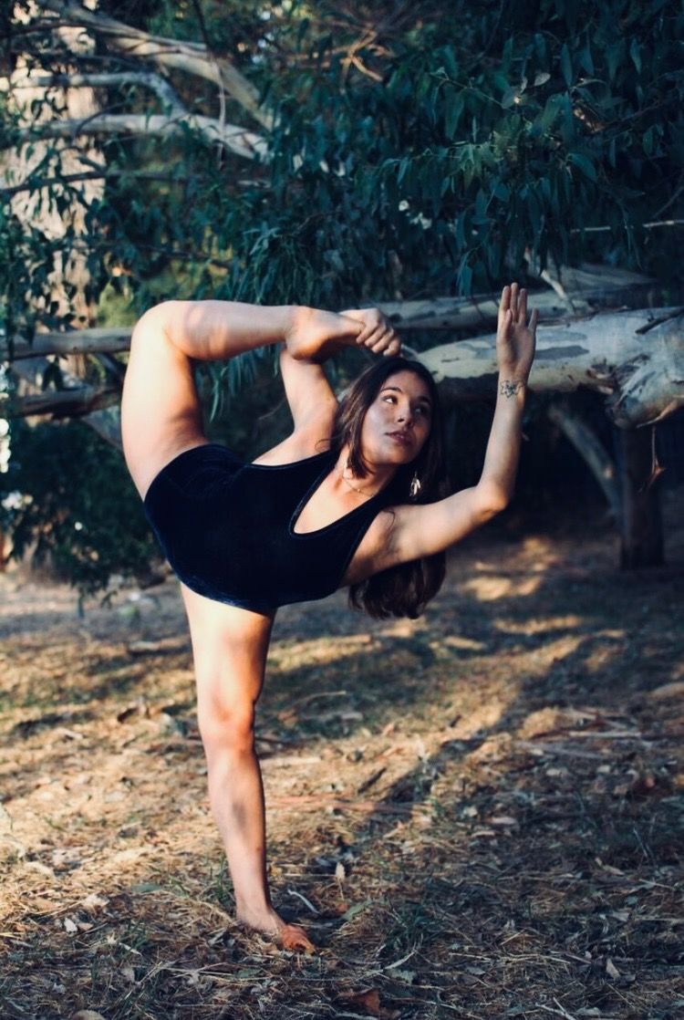 Woman in a black leotard balances in yoga pose outdoors, raising one arm, foot on head, near trees.