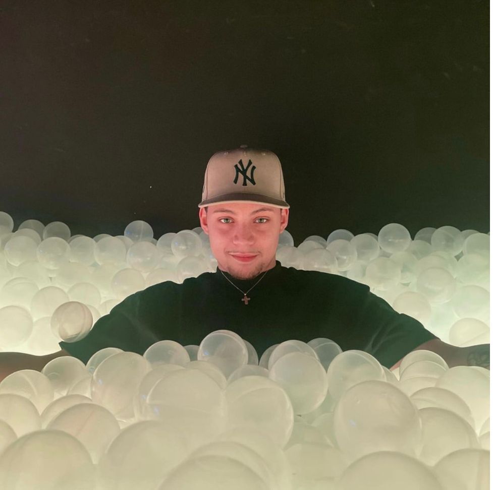 Man in a baseball cap smiles, surrounded by white balls in a dark setting.