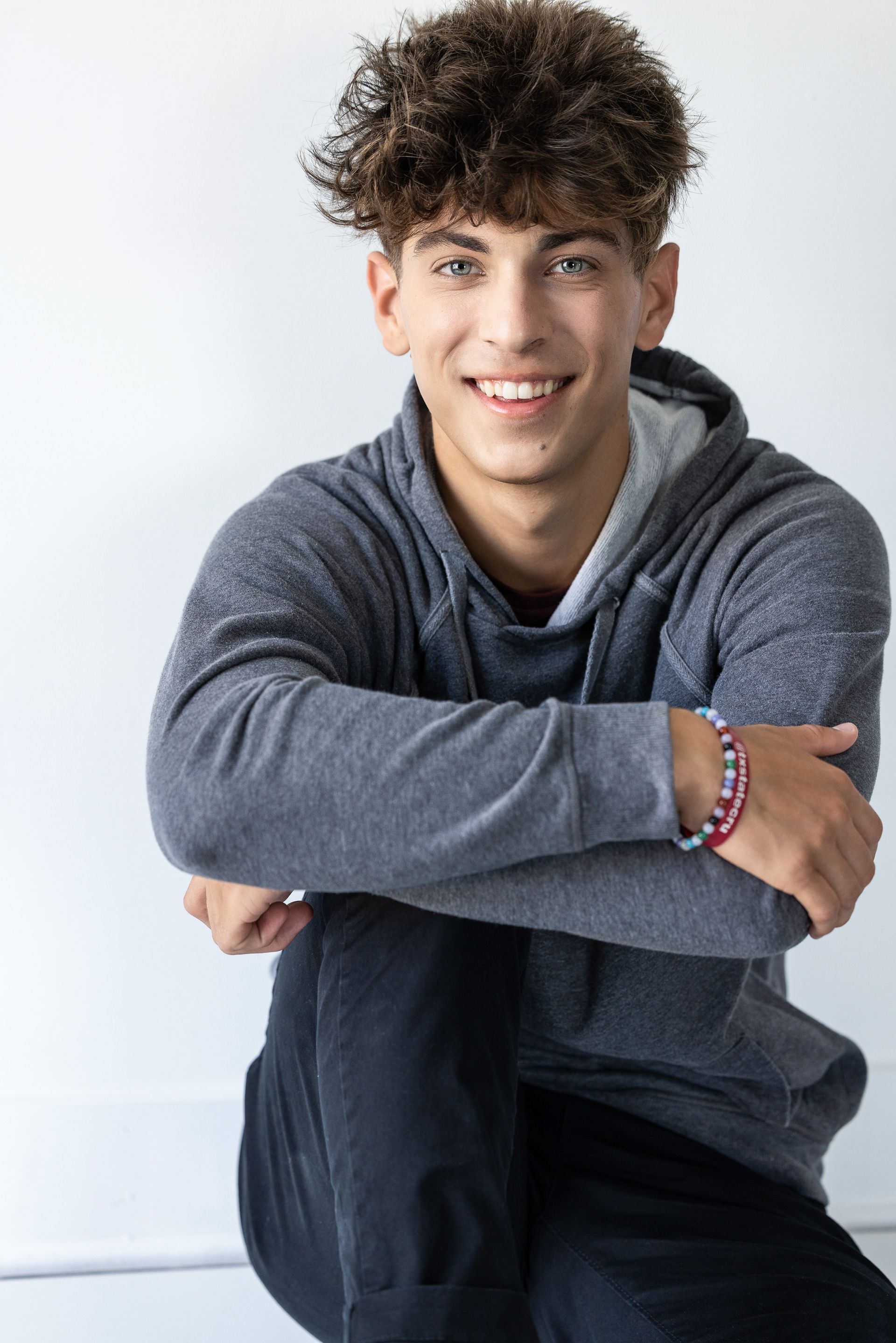 Young person with curly brown hair wearing a gray hoodie, smiling, sitting.