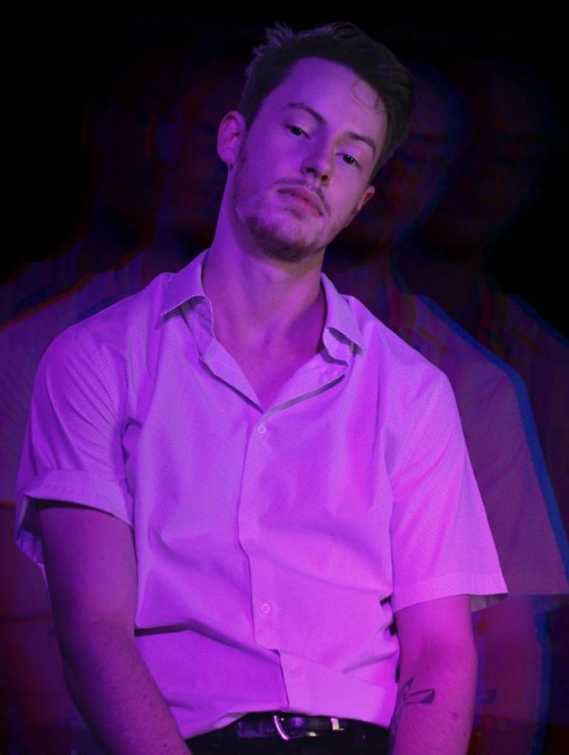 Man in pink shirt, looking at the camera. Purple lighting with a blurred figure in the background.