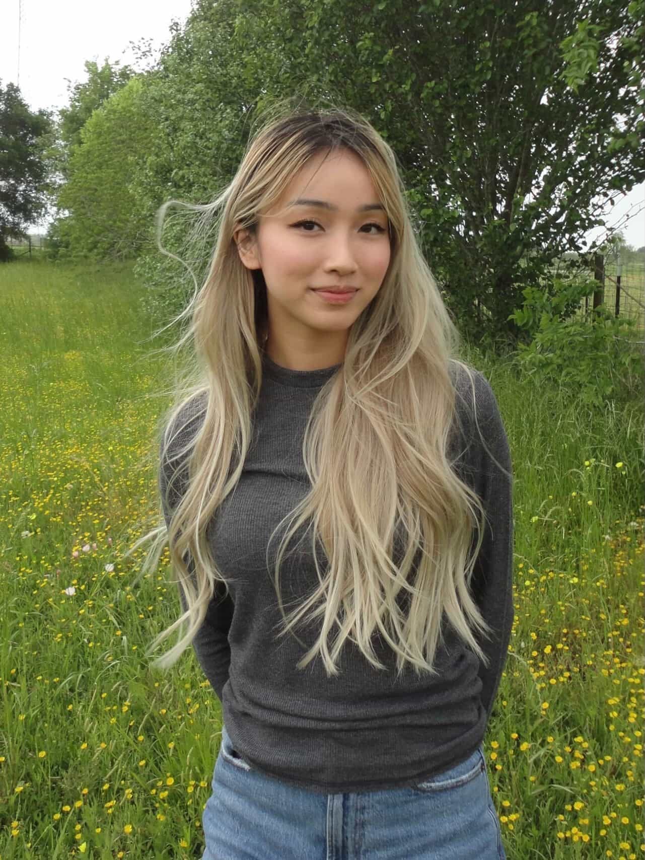 Woman with long blonde hair in a field of yellow flowers, wearing a gray sweater and jeans.
