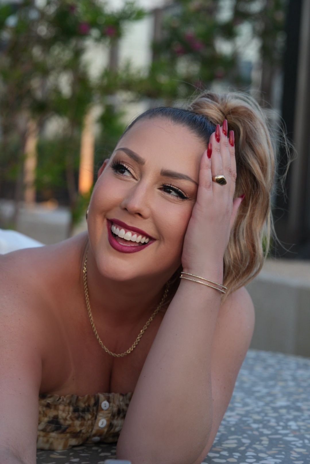 Woman with blonde hair up, smiling, wearing gold jewelry, hand on head, outdoor setting.