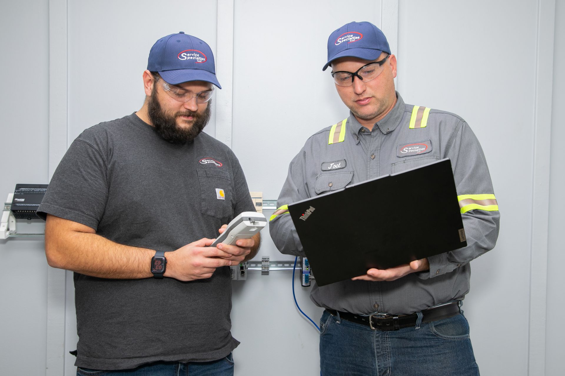 Two technicians in blue caps review a clipboard and phone in a workshop.