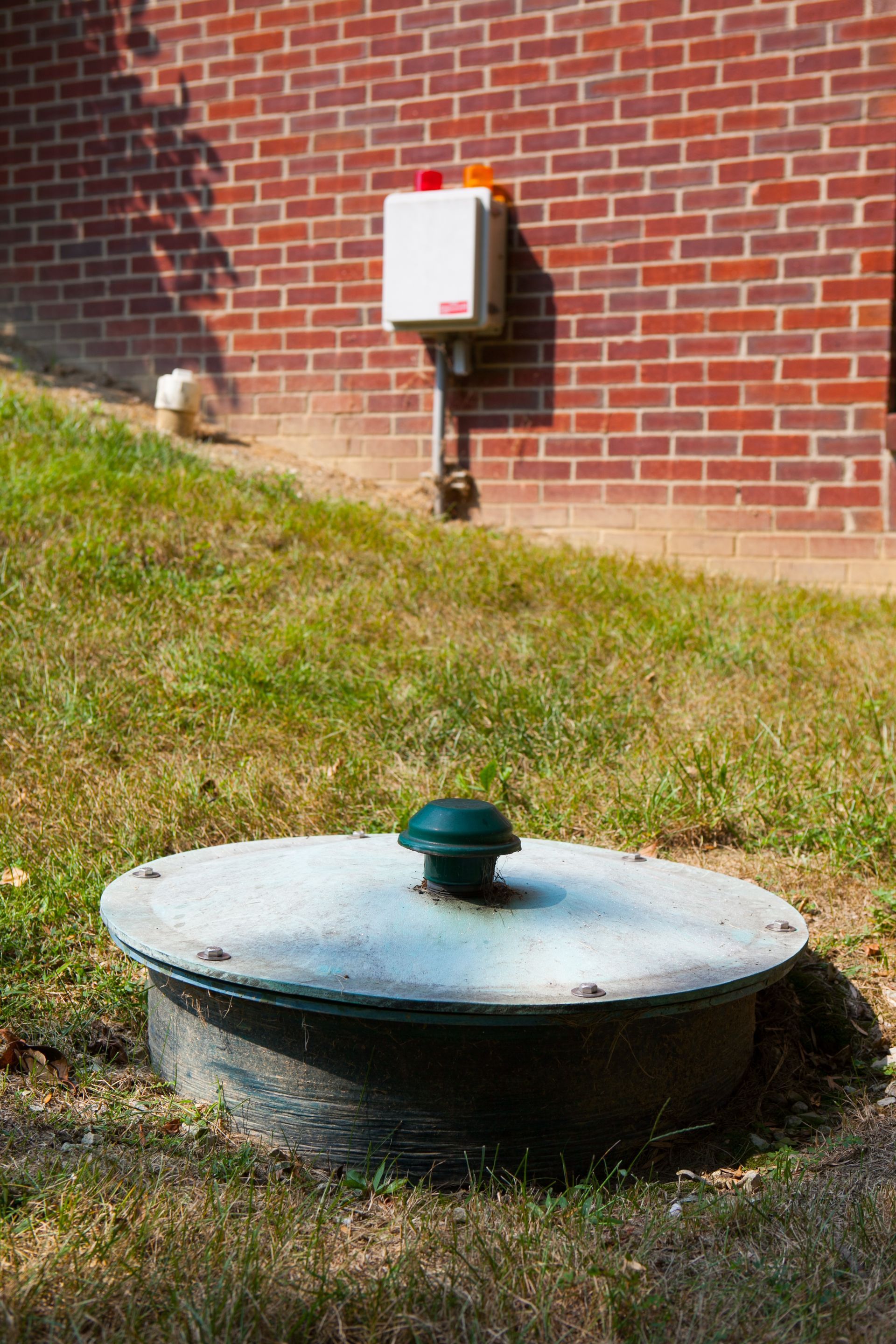 Round utility manhole cover in grassy yard beside a brick building with a wall-mounted electrical box.