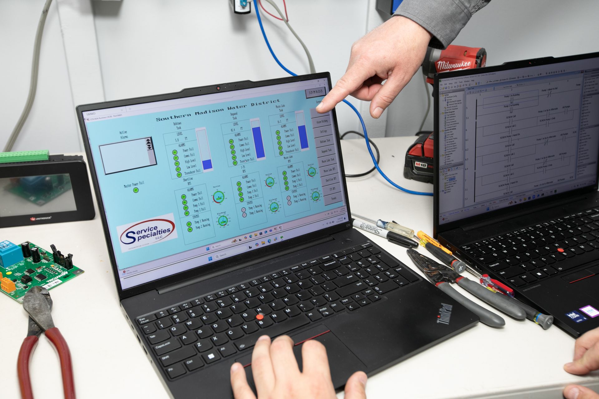 Two laptops displaying technical schematics, with a hand pointing at the left screen on a workbench