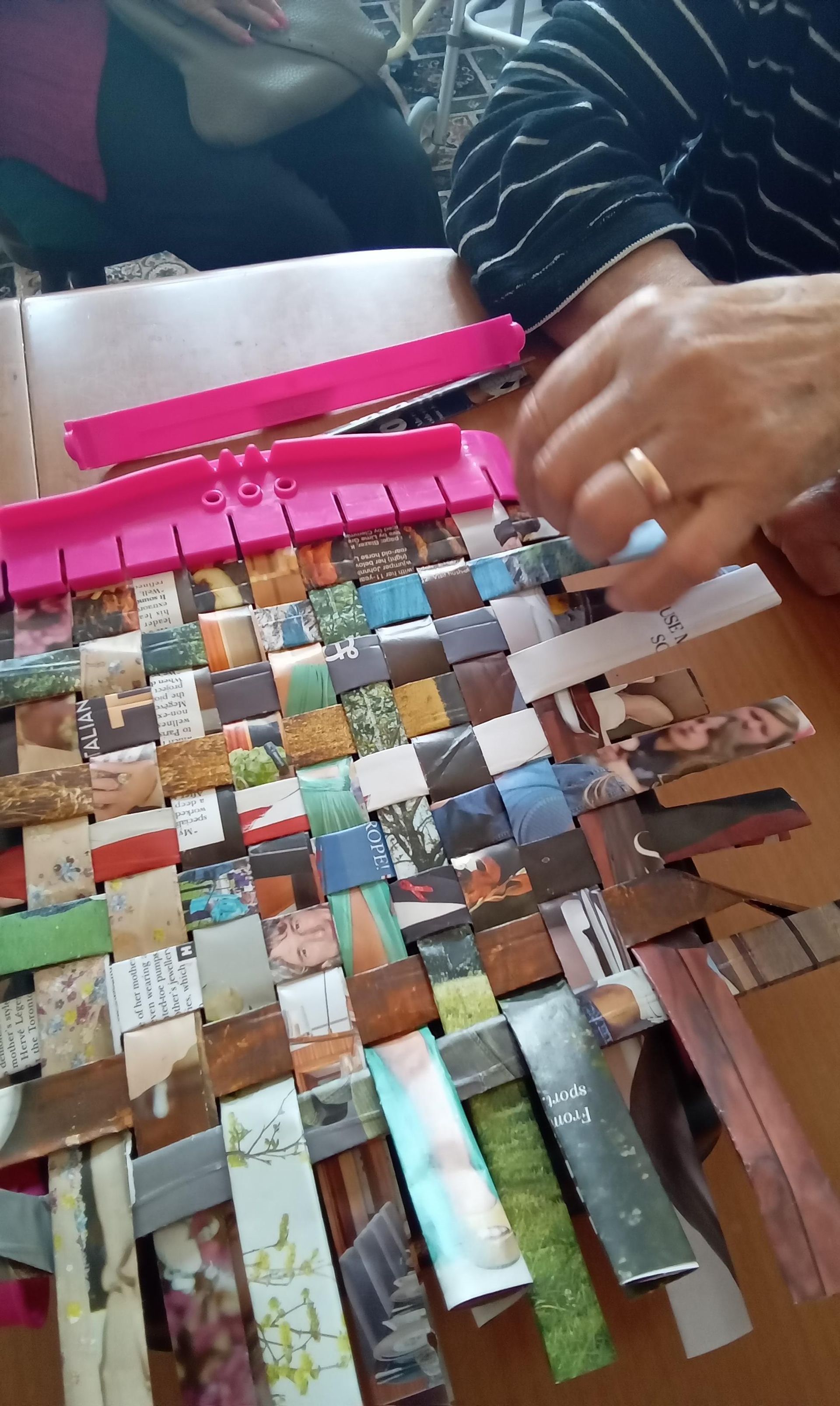 Person weaving strips of magazine paper using a pink plastic weaving loom on a table.