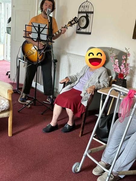 A musician plays guitar for a person in a chair; a walker is nearby. Indoors with decorative elements.