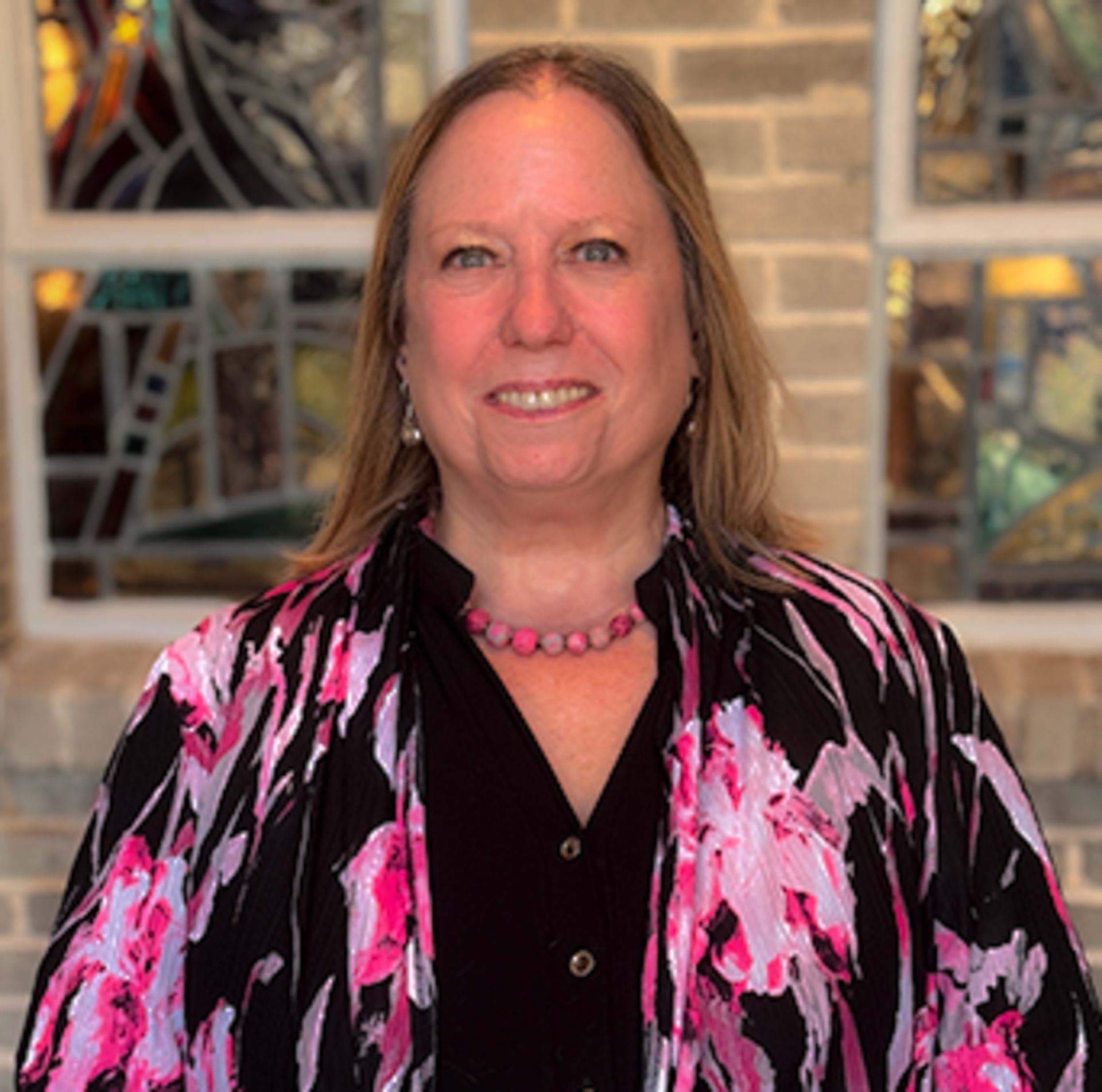 A woman wearing a purple sweater and glasses is smiling in front of a brick wall.