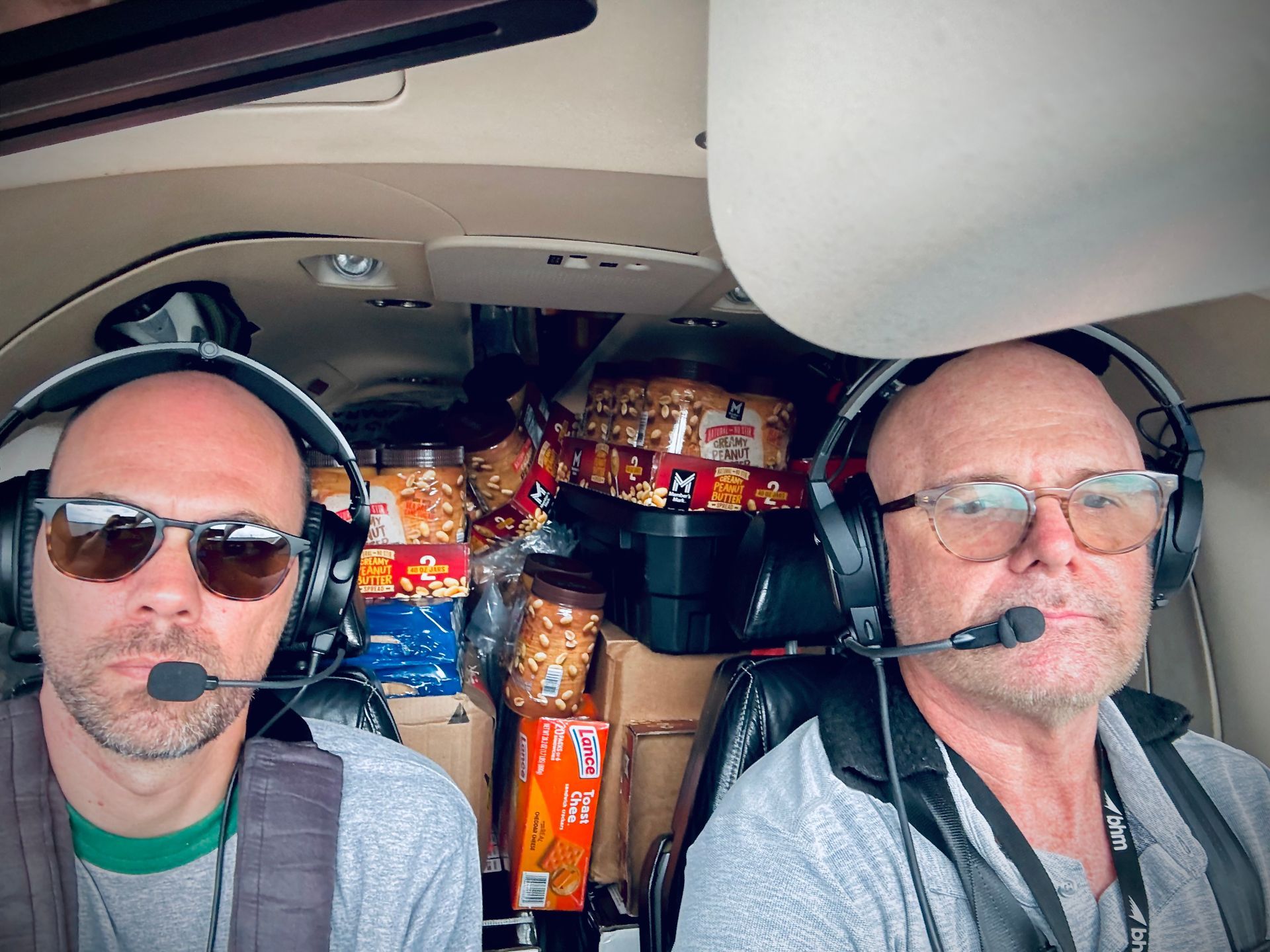 Two men wearing headphones and sunglasses are sitting in a plane.