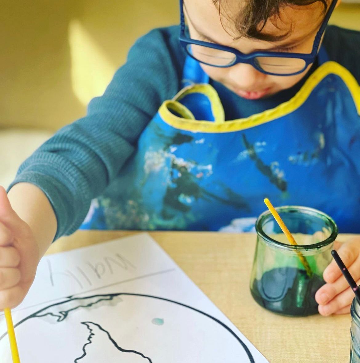 A young boy wearing glasses is drawing a globe on a piece of paper