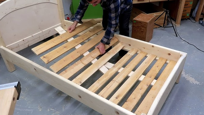 A man is working on a wooden bed frame in a workshop.