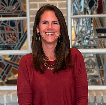 A woman in a red shirt is smiling in front of a stained glass window.