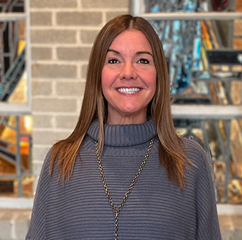 A woman wearing a sweater and a necklace is smiling in front of a stained glass window.