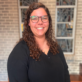 A woman wearing glasses and a black shirt is smiling in front of a brick wall.